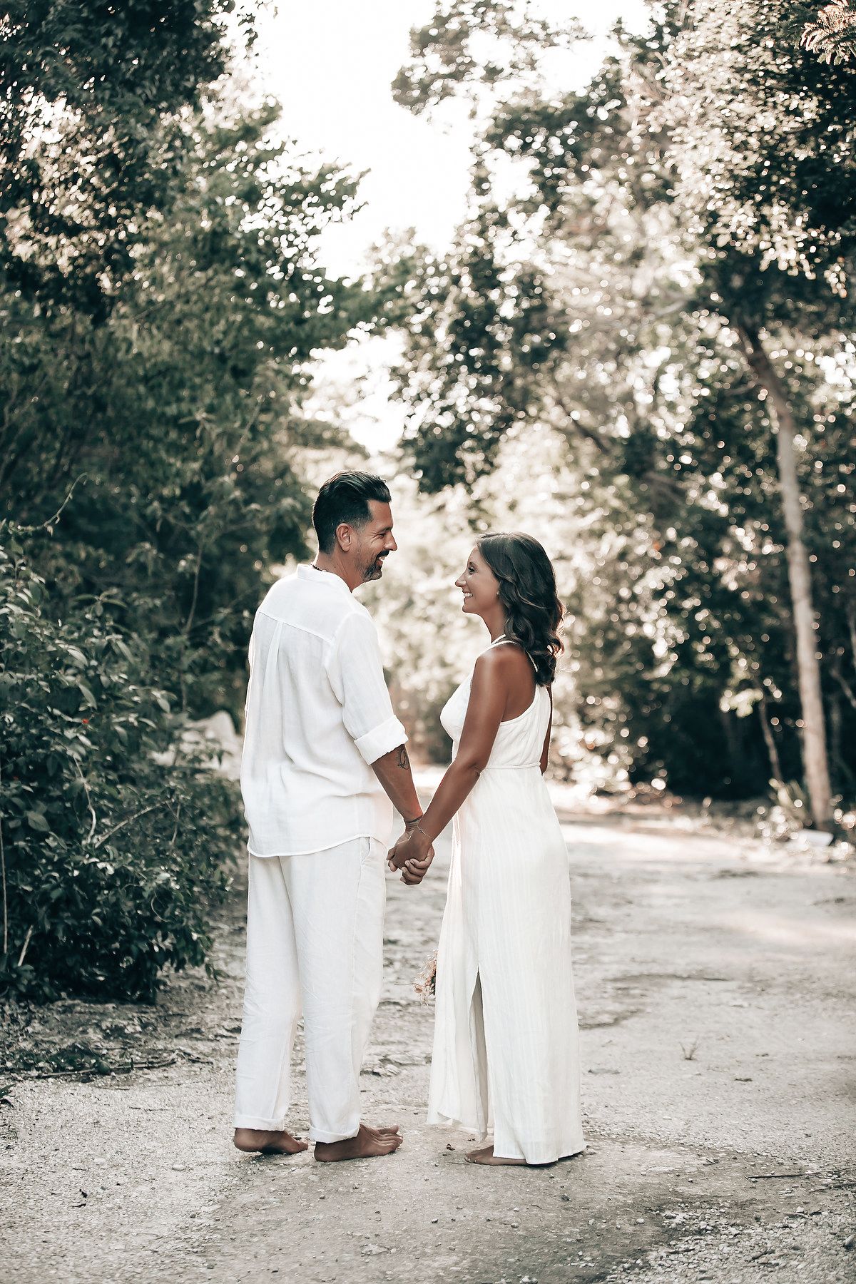 A couple stands together in a lush, forested landscape, surrounded by greenery and a body of water.