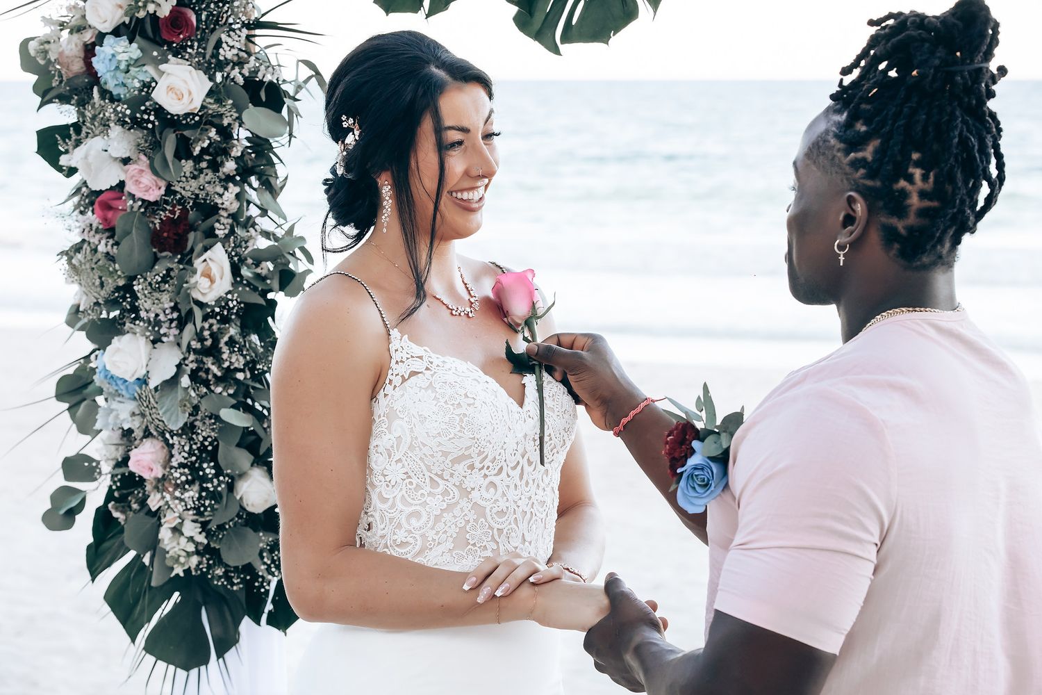 A woman in a pink outfit stands in front of a floral arch on a beach.