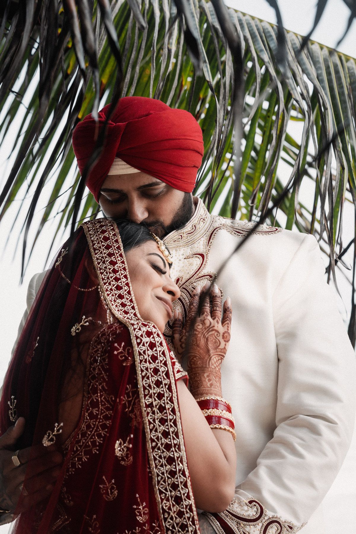 Sikh groom in red turban tenderly embraces bride in red lehenga under tropical palm leaves.