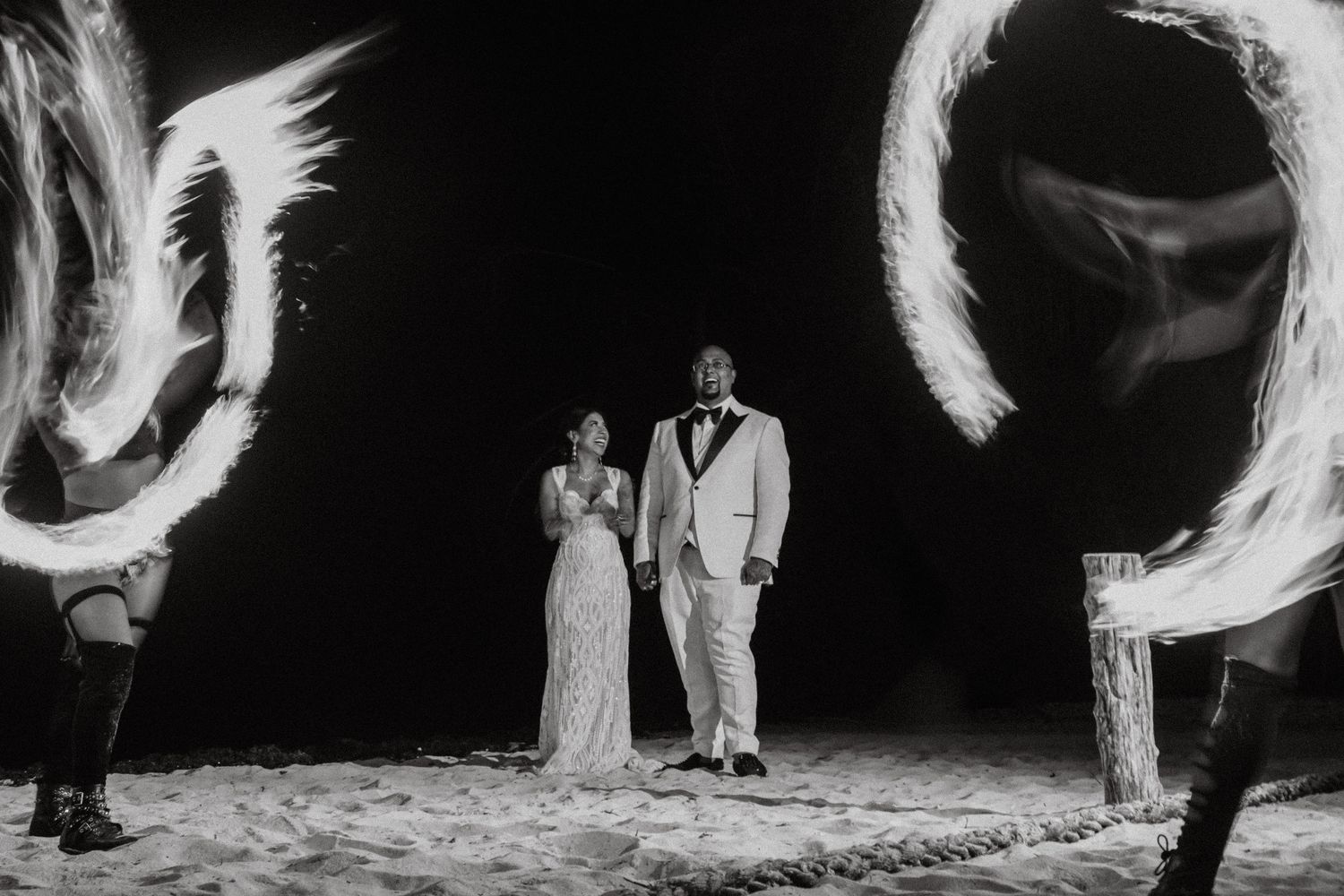 Black and white photo of bride and groom walking through fire dancers on beach at night wedding.