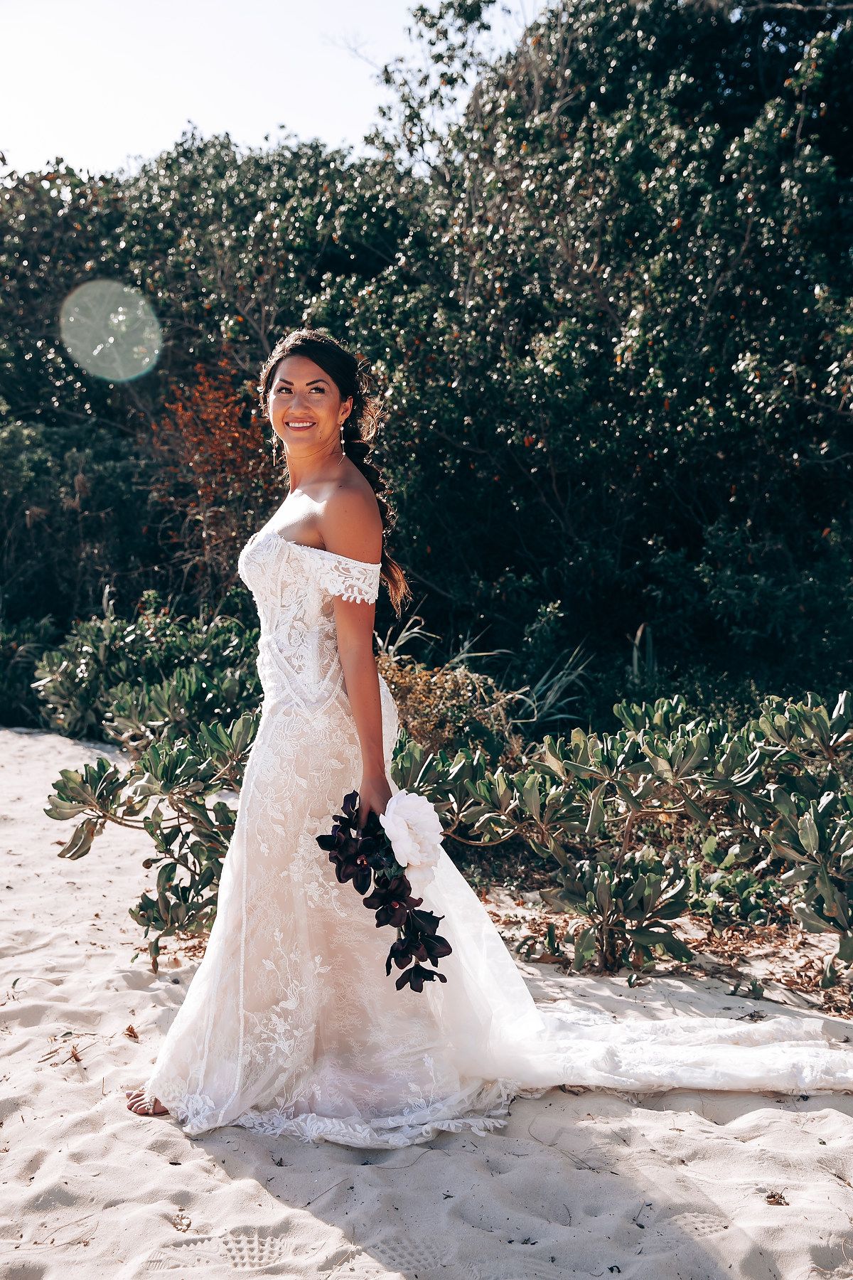 White lace wedding dress displayed against tropical foliage on a sandy beach.