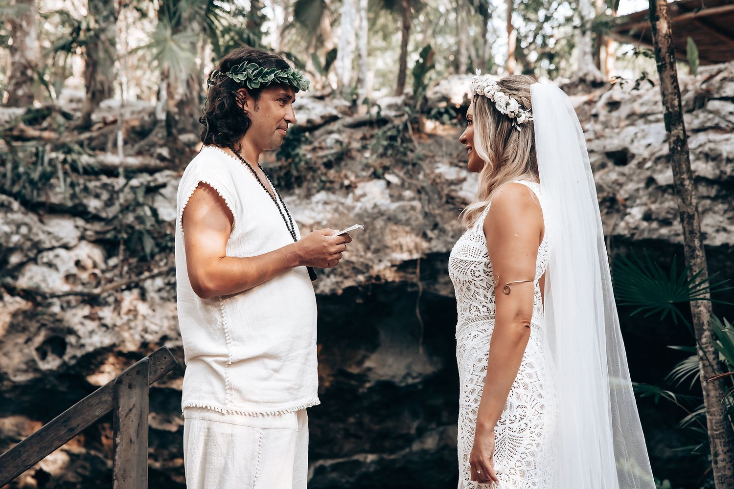 A couple in white attire stands together during a wedding ceremony in front of a rocky cliff backdrop.