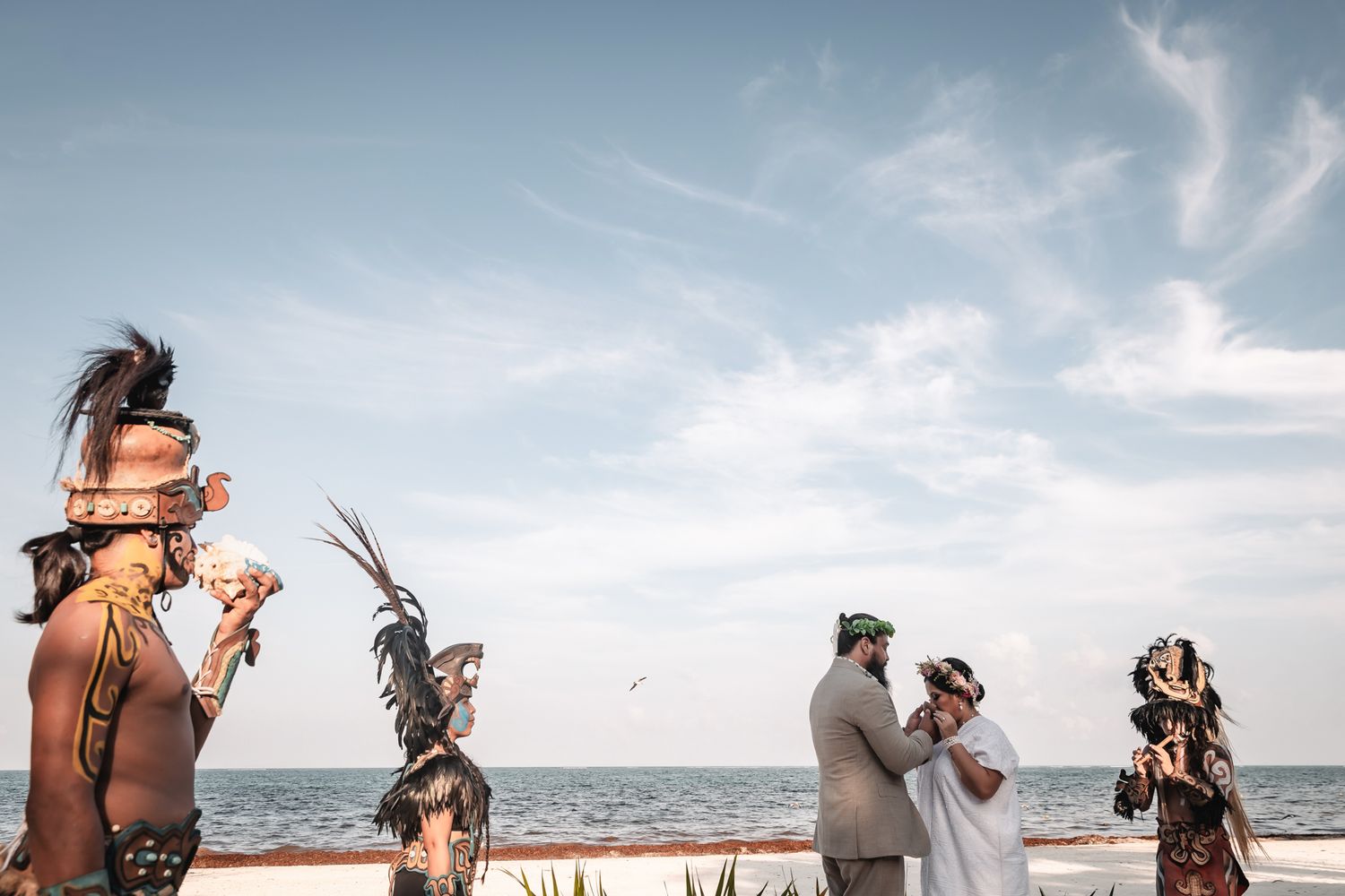 Indigenous performers in traditional attire conduct a beach ceremony near the ocean under a partly cloudy sky.