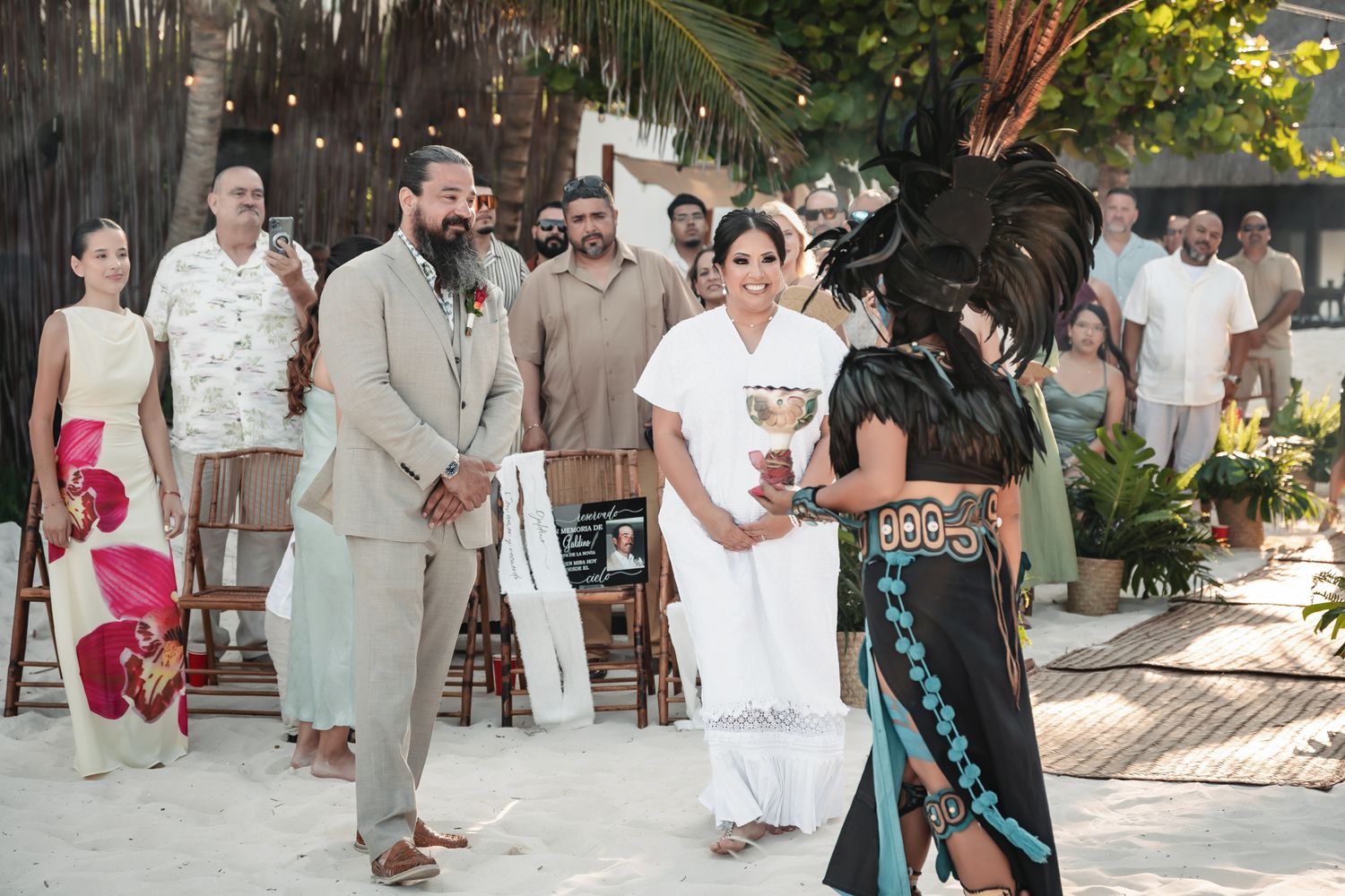 Couple stands before Mayan performer in traditional headdress during outdoor beach wedding ceremony with guests watching.