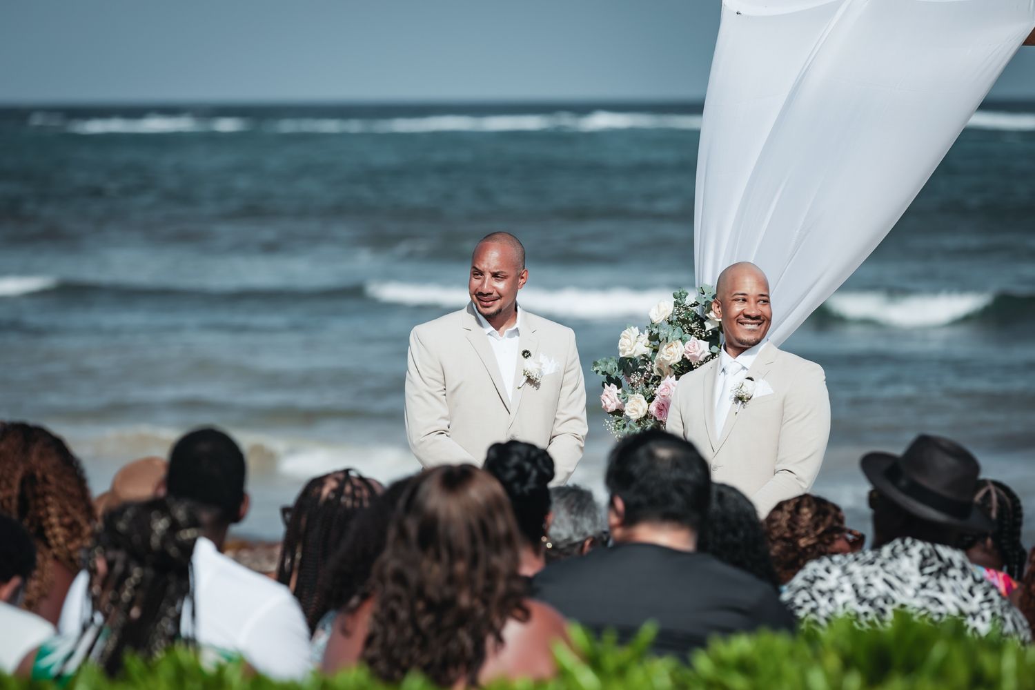 Two grooms in cream suits smile at beach wedding ceremony with ocean backdrop and white floral arch.