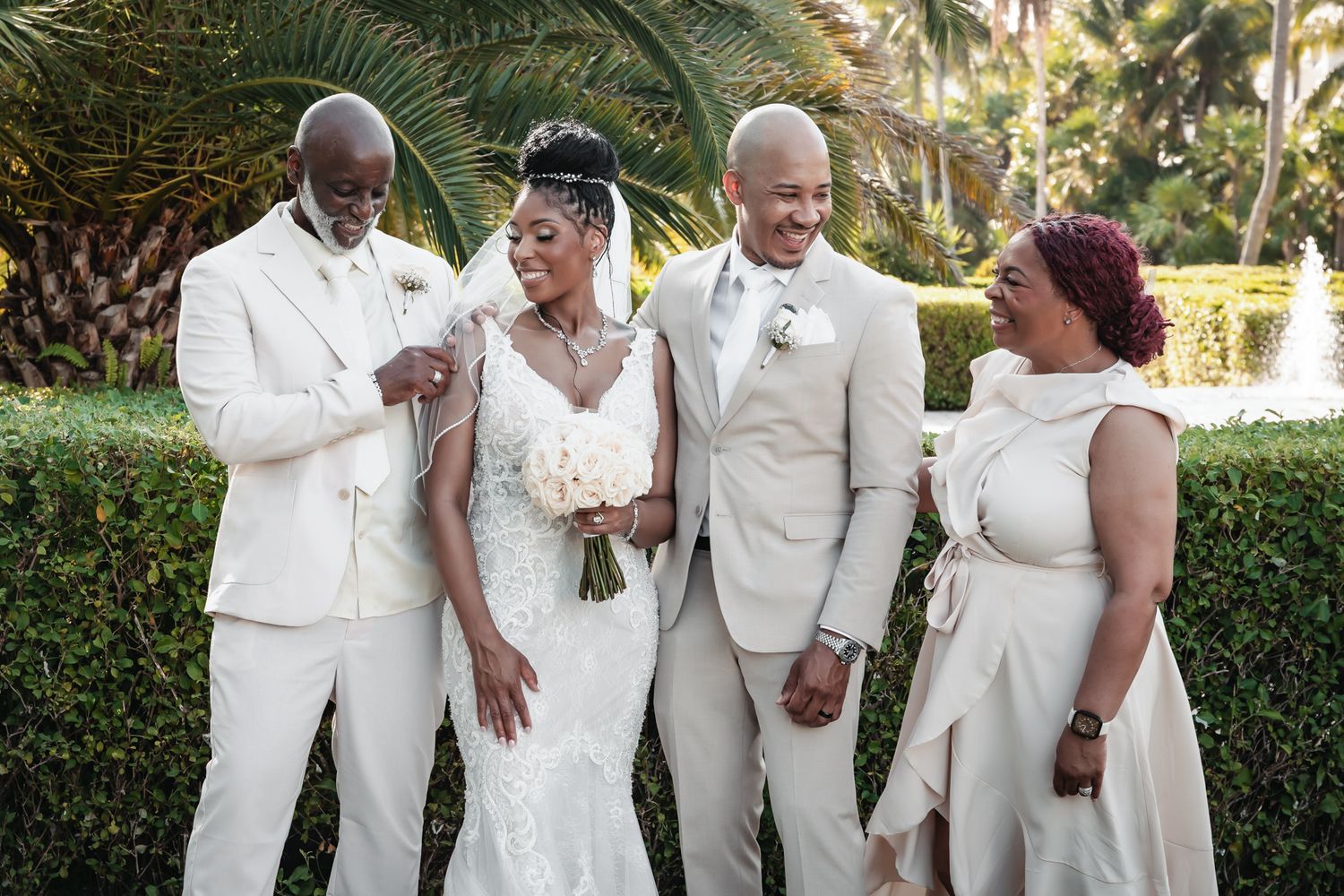 Bride and groom pose with family in elegant white and beige wedding attire surrounded by lush tropical greenery.