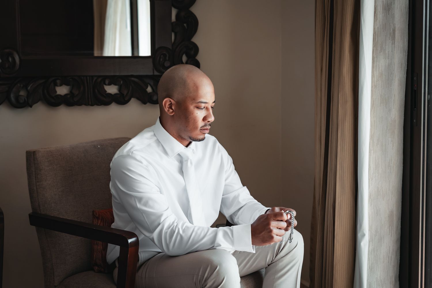 Bald man in white shirt and bow tie sits thoughtfully by window holding a small object in a elegant room.