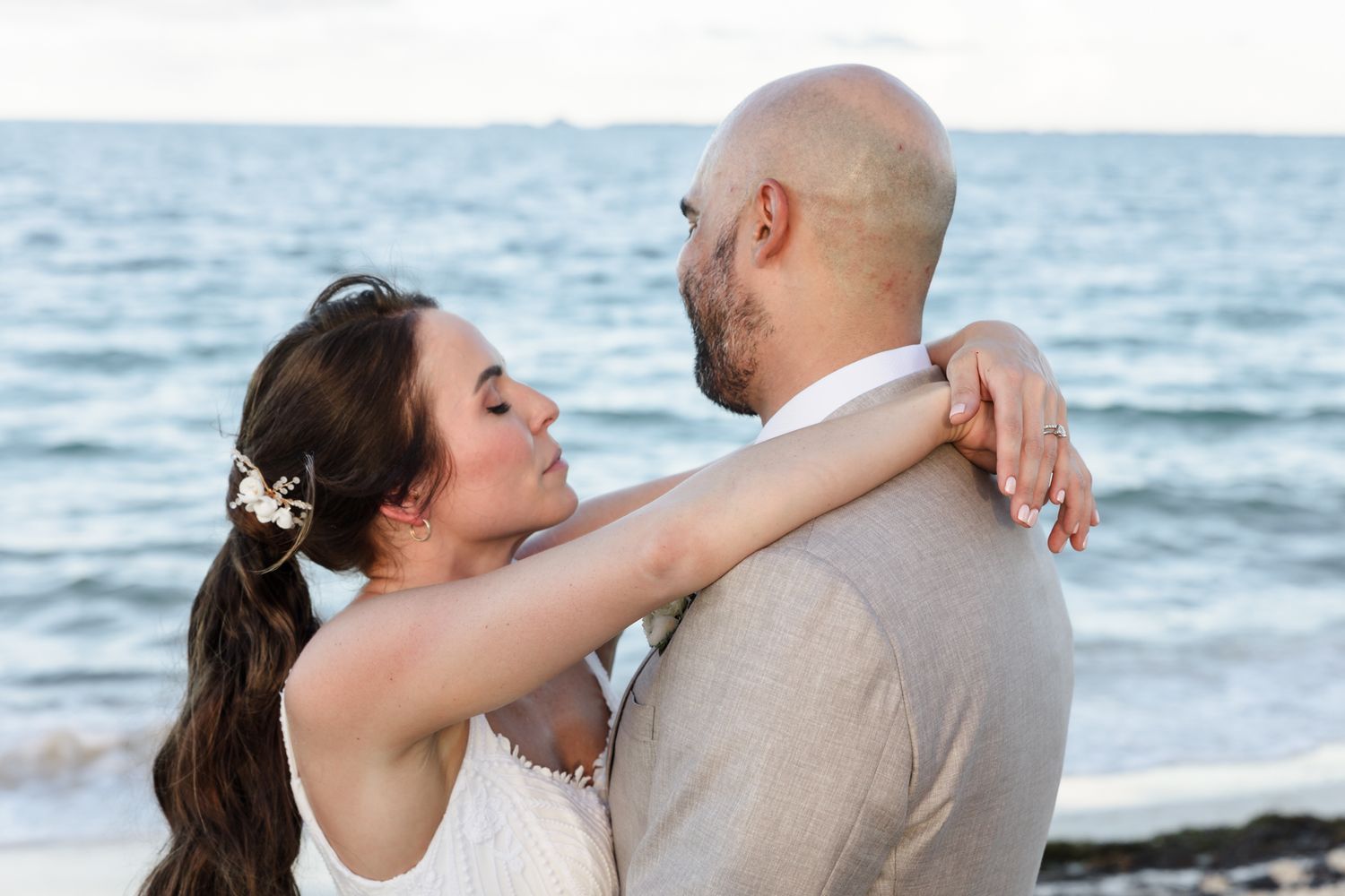 A bride and groom share a romantic embrace on a beach, with ocean waves in the background on their wedding day.