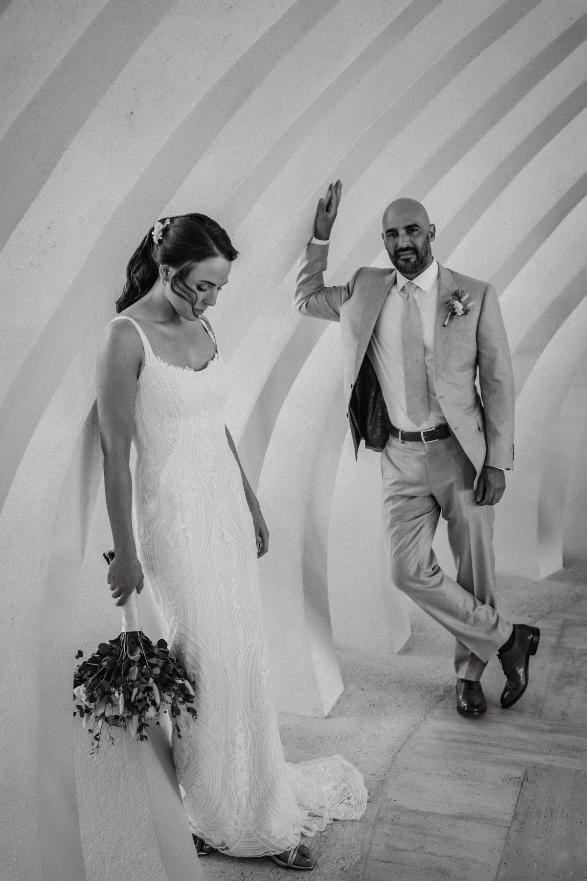 Black and white wedding photo of bride holding bouquet and groom waving on curved staircase.