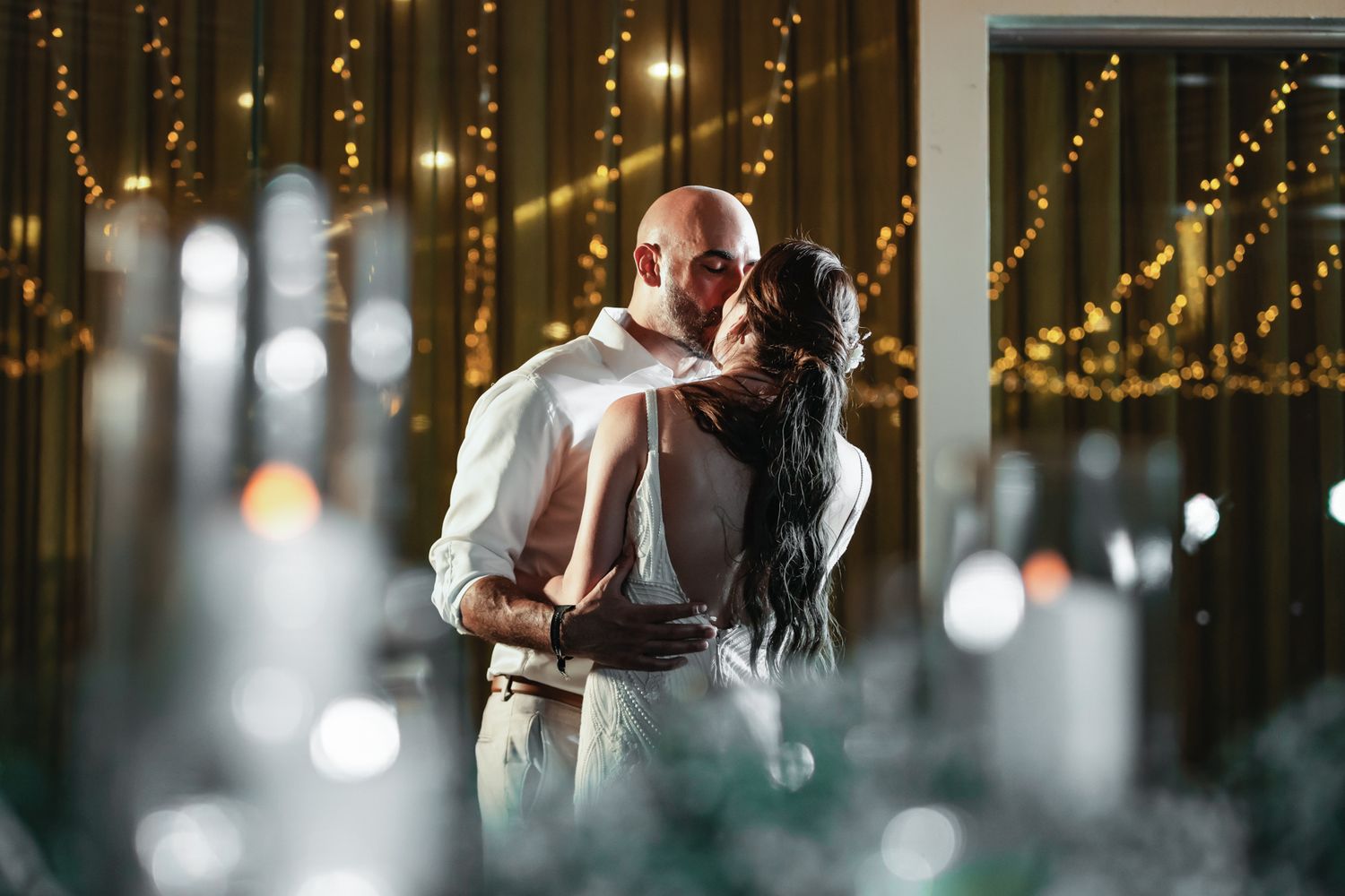 Couple sharing romantic first dance kiss at wedding reception with warm fairy lights and bokeh background.