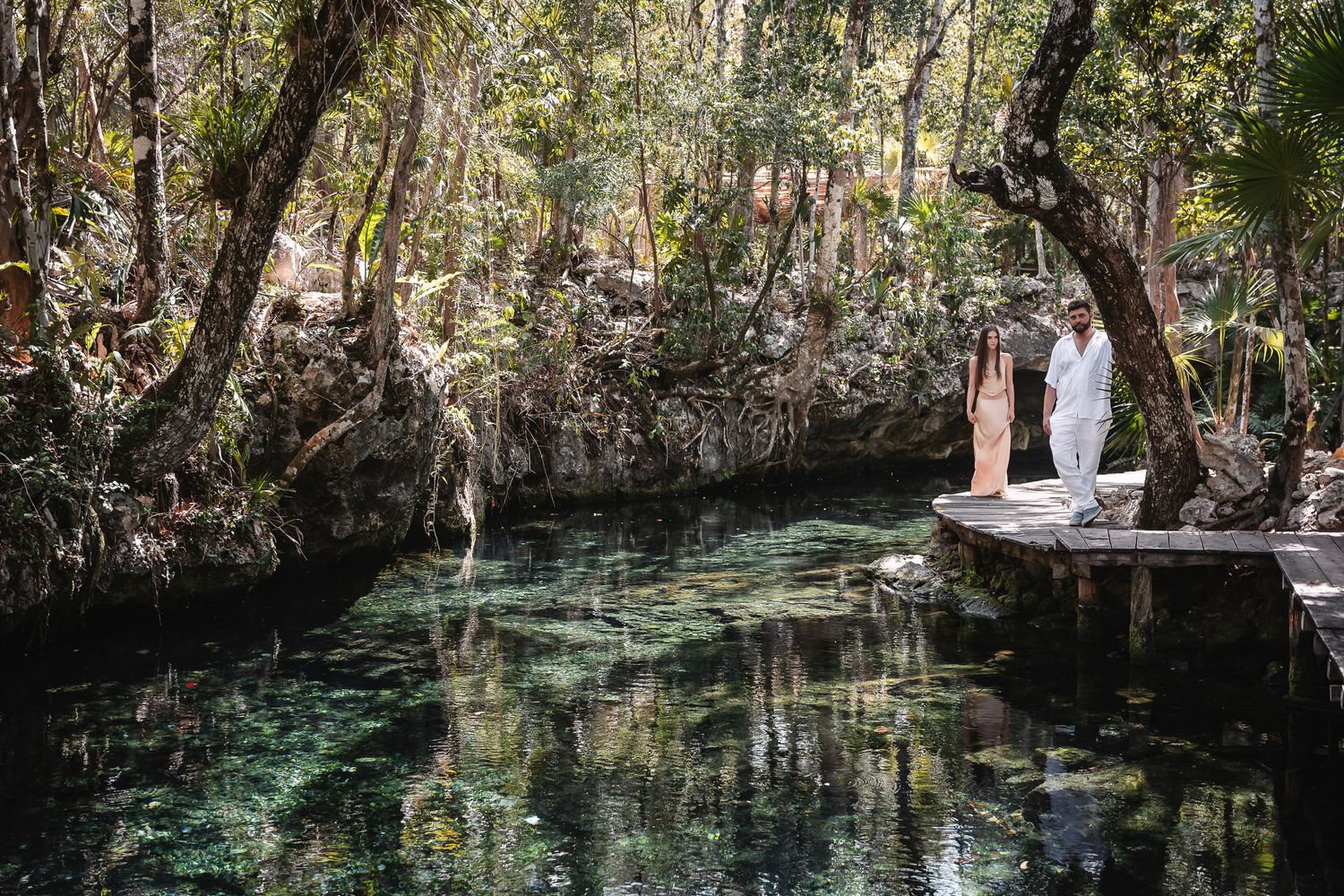 Couple in white attire standing by a serene cenote surrounded by lush jungle trees in Mexico.