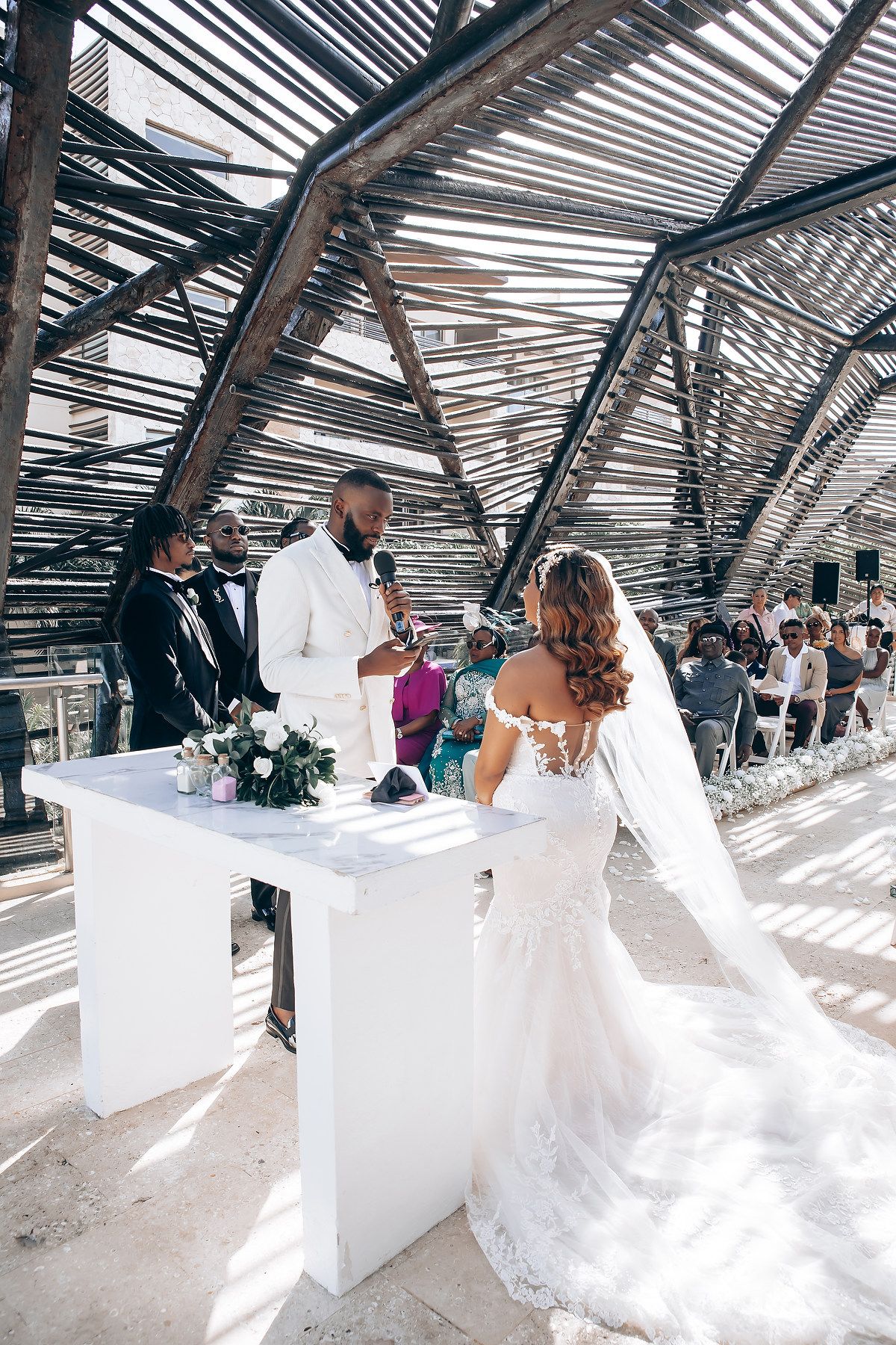 Wedding ceremony setup with white chairs and decor under a modern architectural structure with slatted shadows.