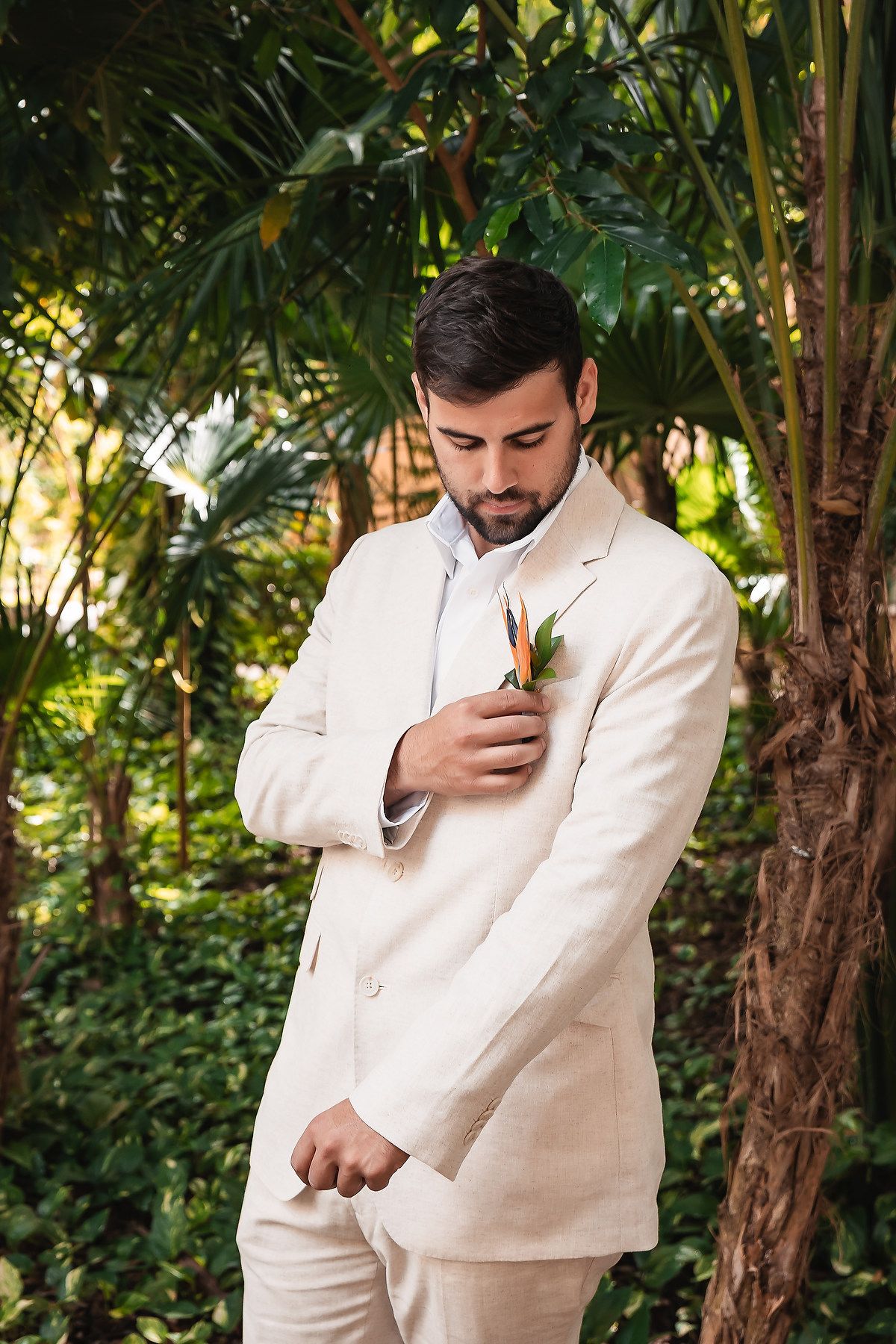 Groom in cream suit adjusts boutonniere in tropical garden setting.