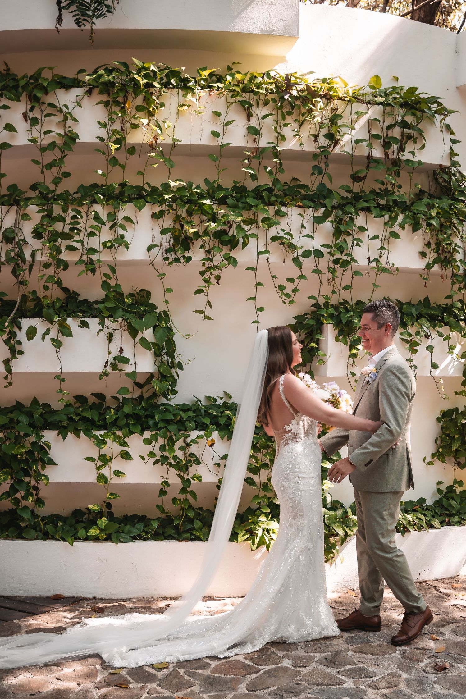 A couple shares a romantic first look moment against a white wall with climbing vines in an outdoor garden setting.