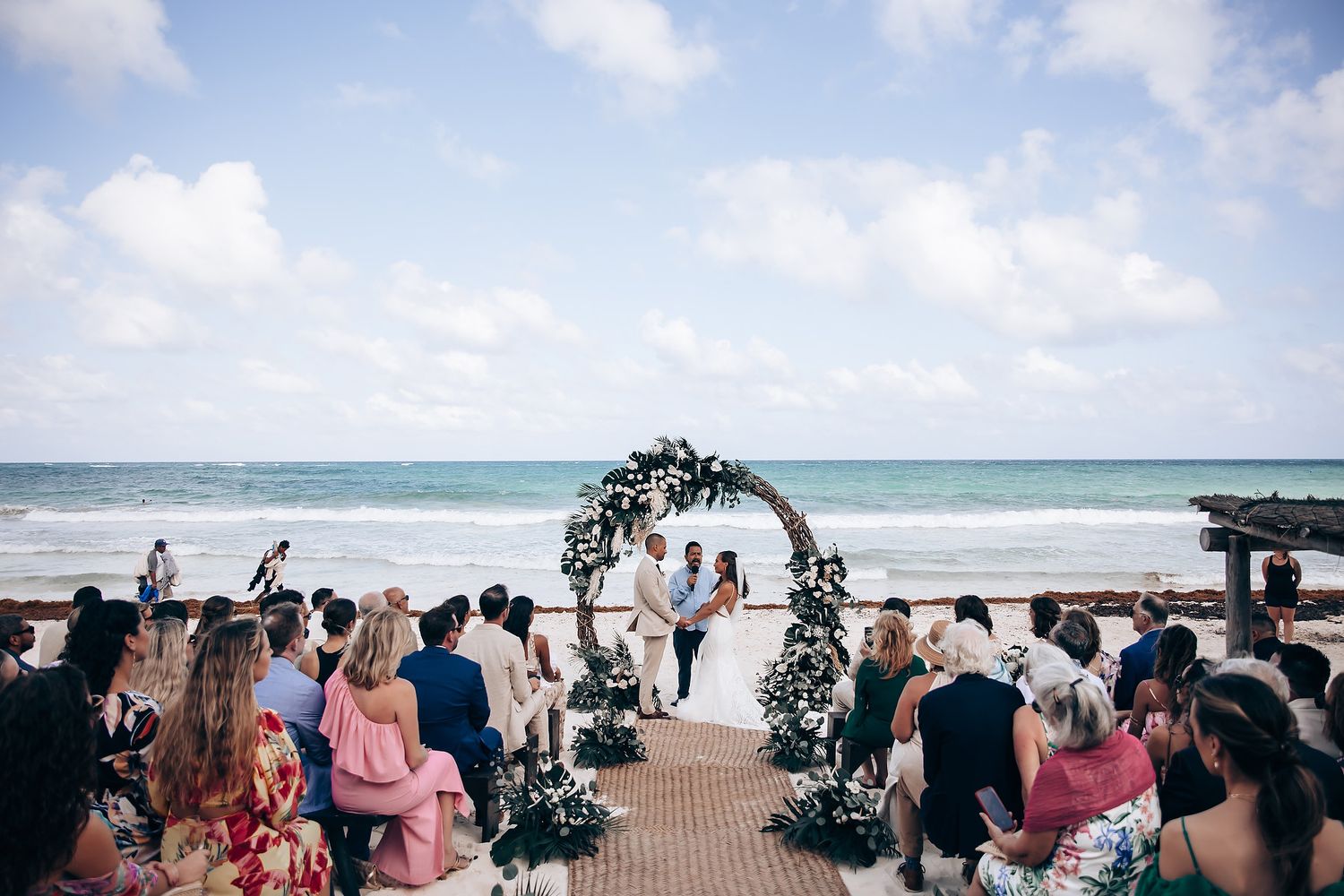 Wide angle shot of a beach wedding ceremony with attendees seated on white chairs.