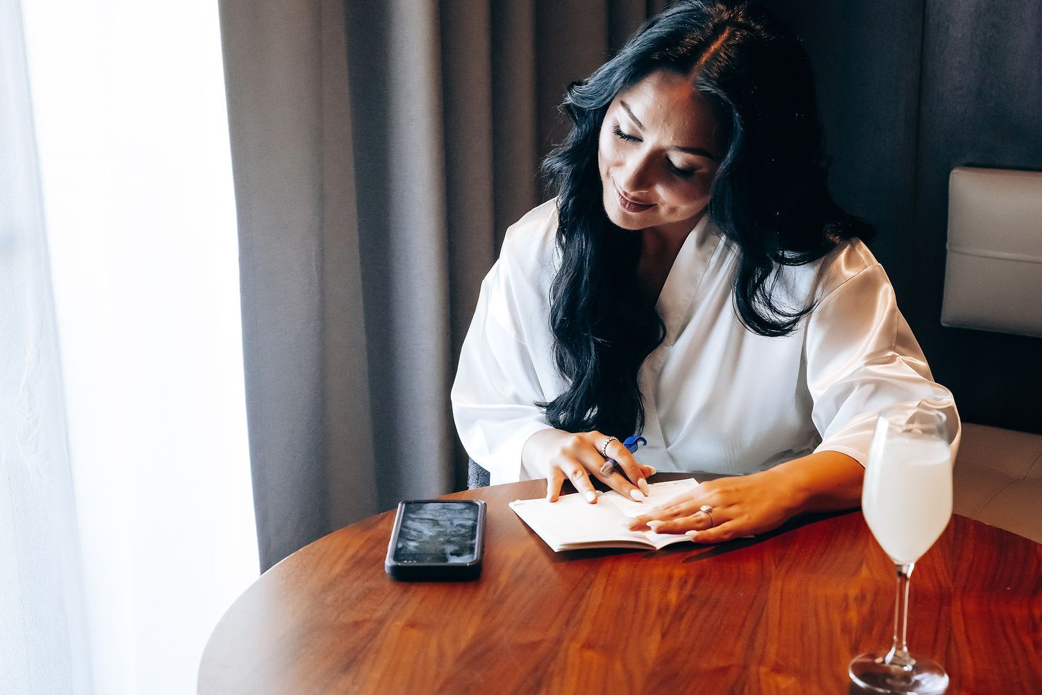 Person in business attire sitting at wooden table with smartphone and glass of white wine.