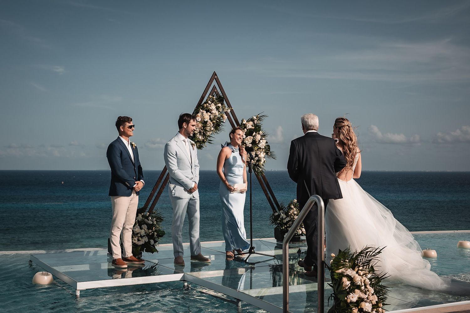 Wedding ceremony in progress under geometric arch with ocean views and floral arrangements.