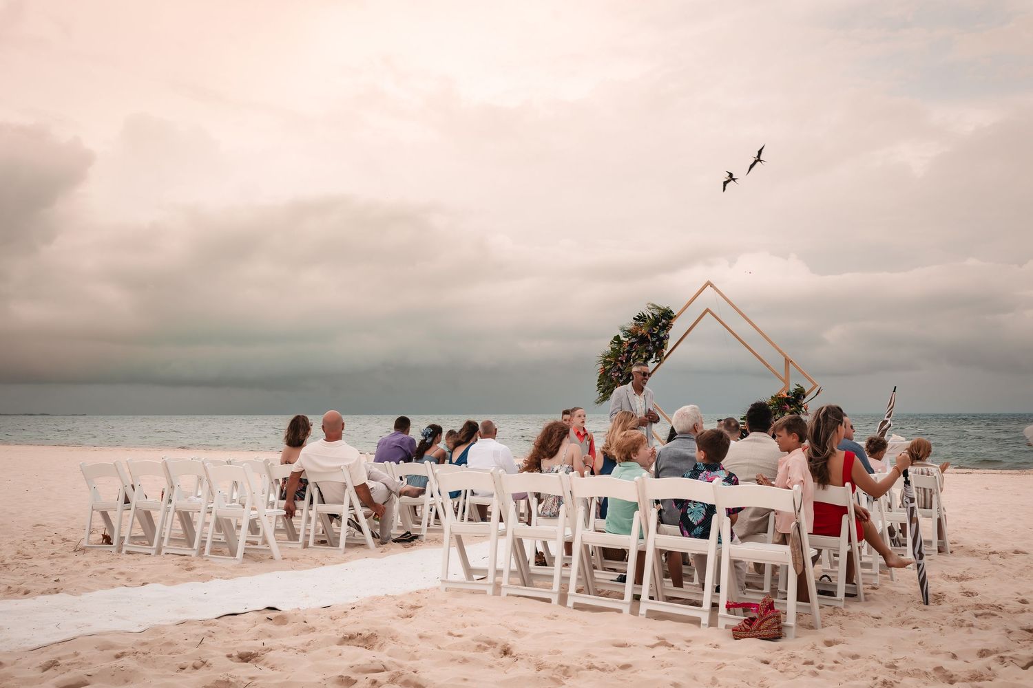 Beach wedding ceremony setup with white chairs and wooden arch against a cloudy ocean backdrop.