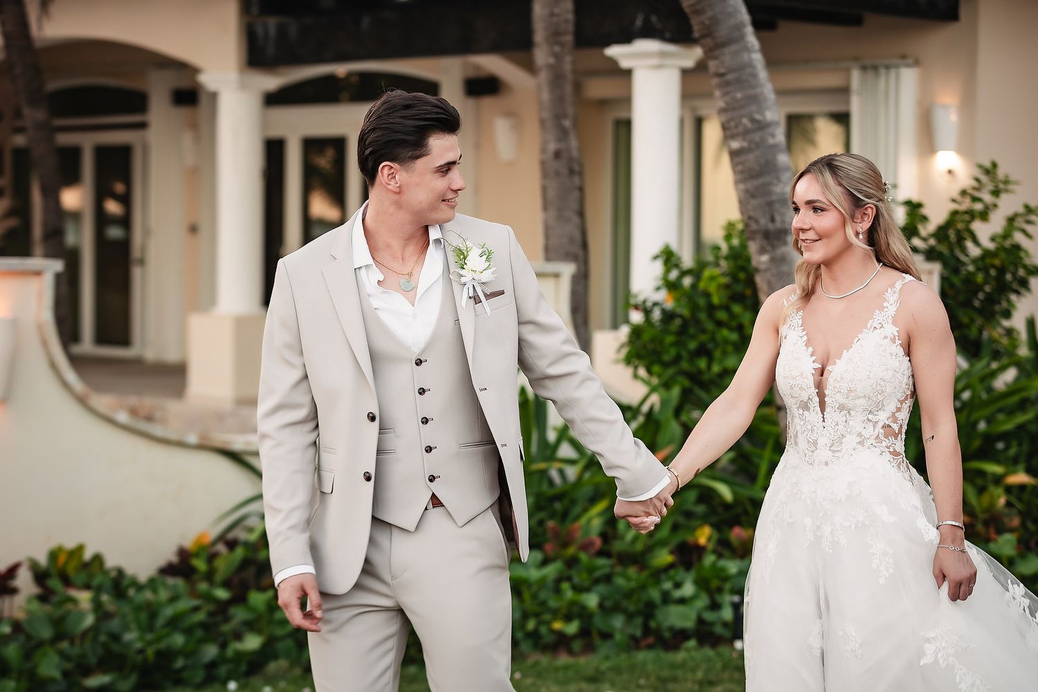 Wedding couple in light gray suit and lace gown stroll through lush garden setting.