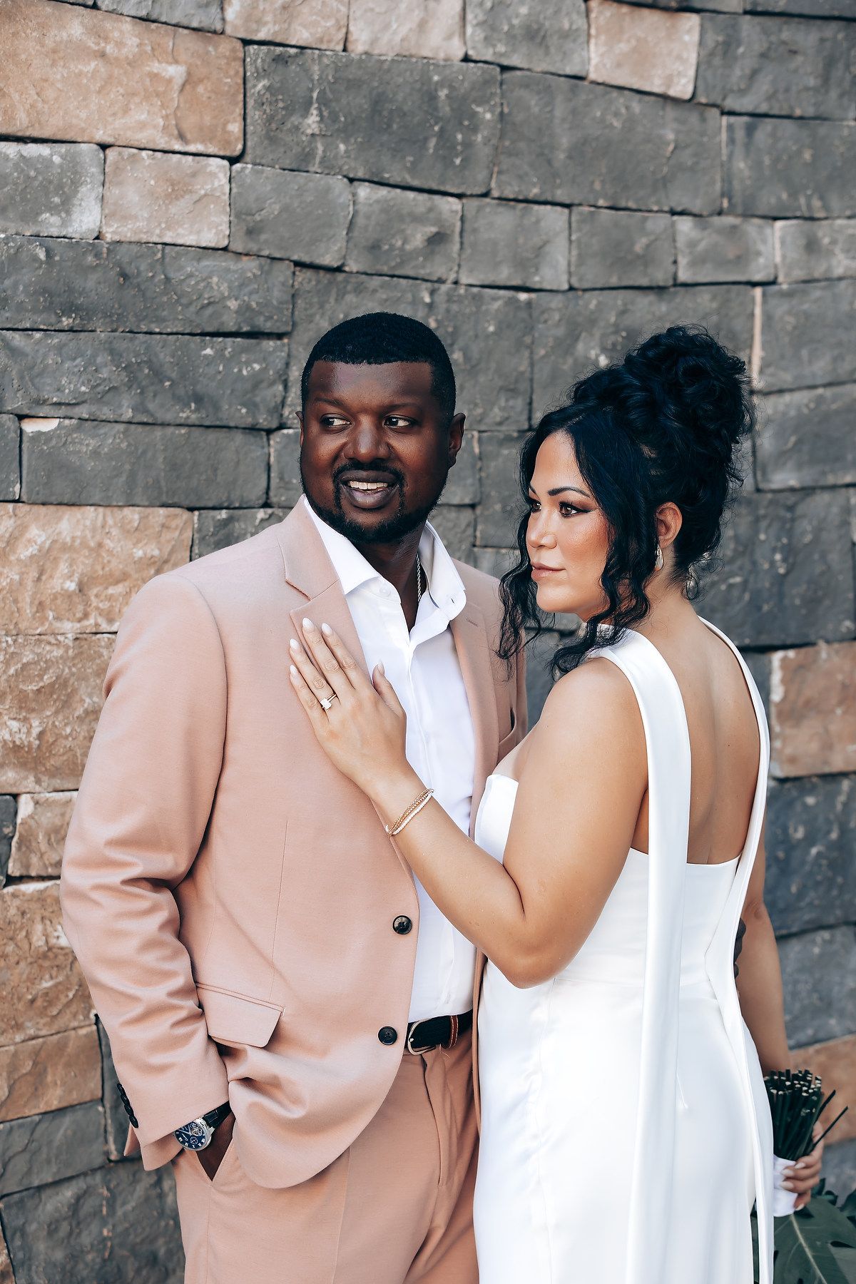 A couple poses against a stone wall wearing coordinated blush pink and white formal attire.