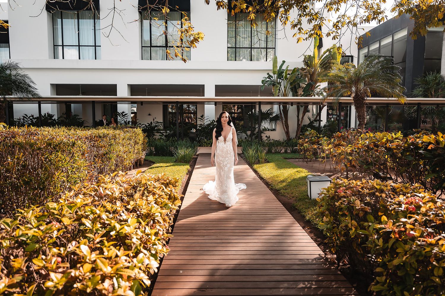 A bride walks along a wooden pathway surrounded by lush yellow fall gardens at a luxury venue.