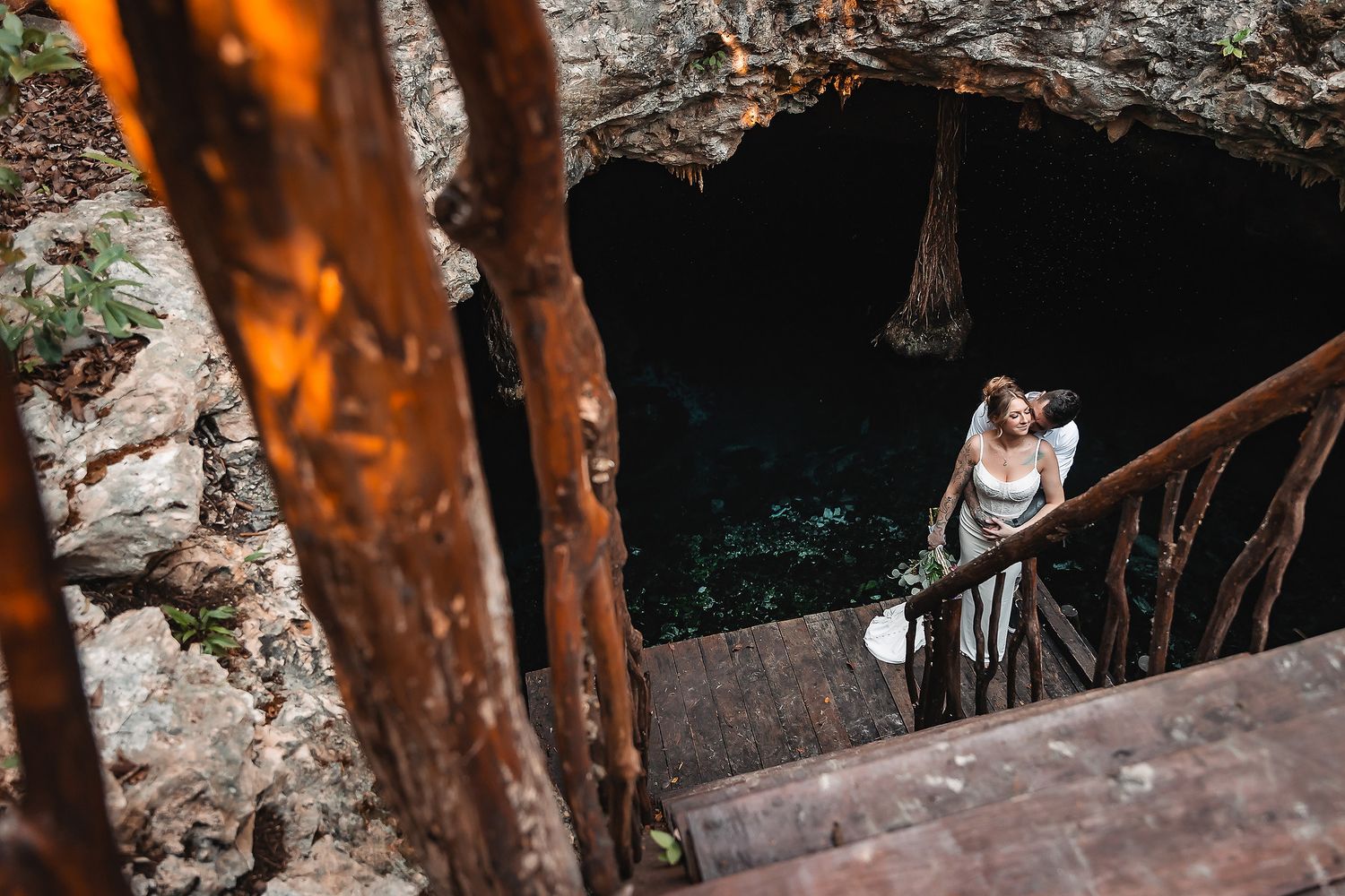 A couple stands on wooden stairs leading down to a cenote cave with crystal clear turquoise water in Mexico.