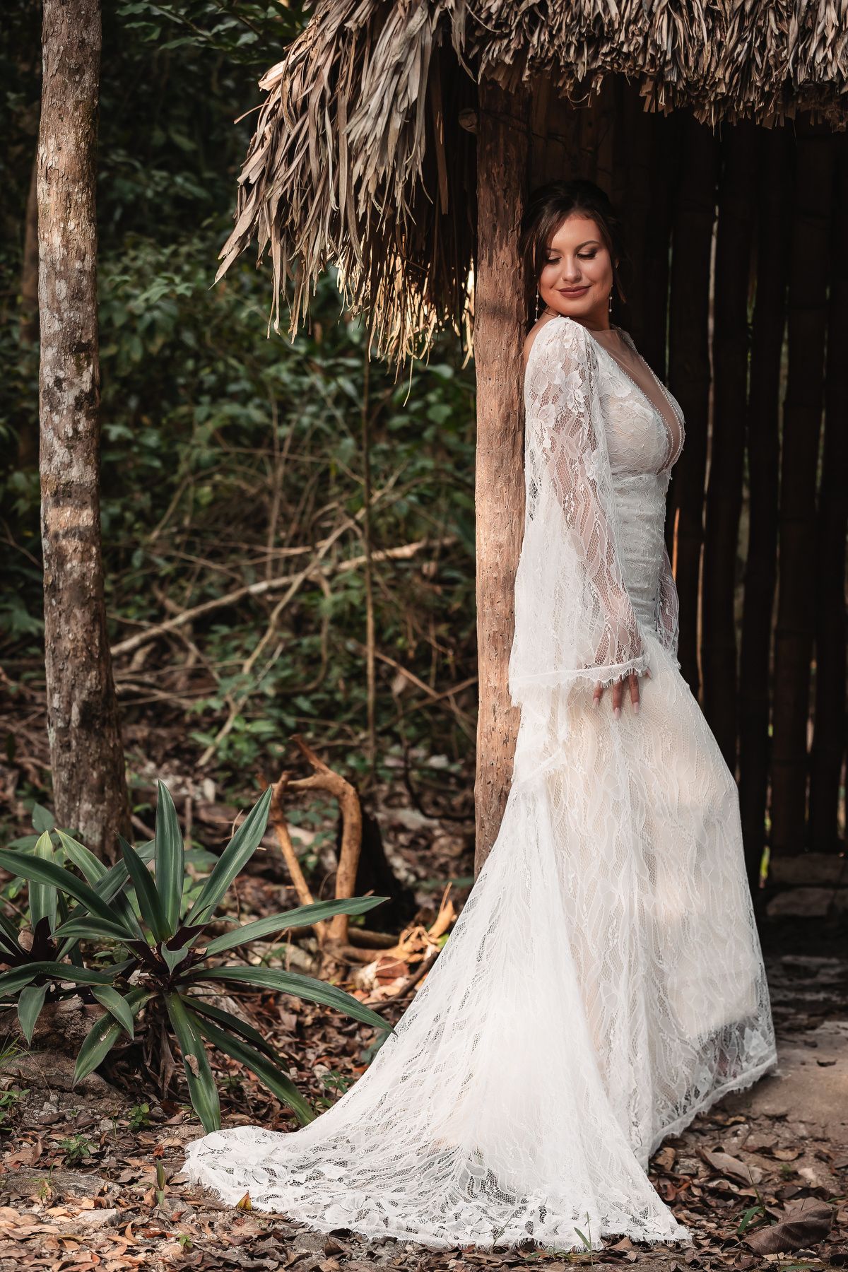 A bride in a form-fitting lace wedding dress poses against a rustic thatched-roof structure.