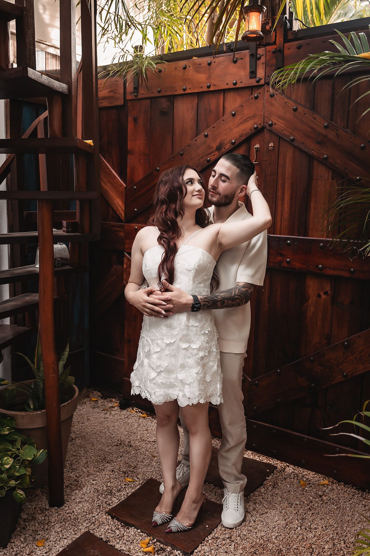 A couple shares an intimate moment against a rustic wooden fence backdrop with tropical plants.