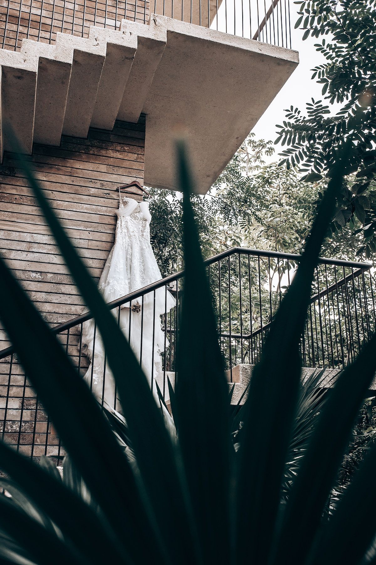 A white wedding dress hangs elegantly against a brick wall with tropical foliage in the foreground.