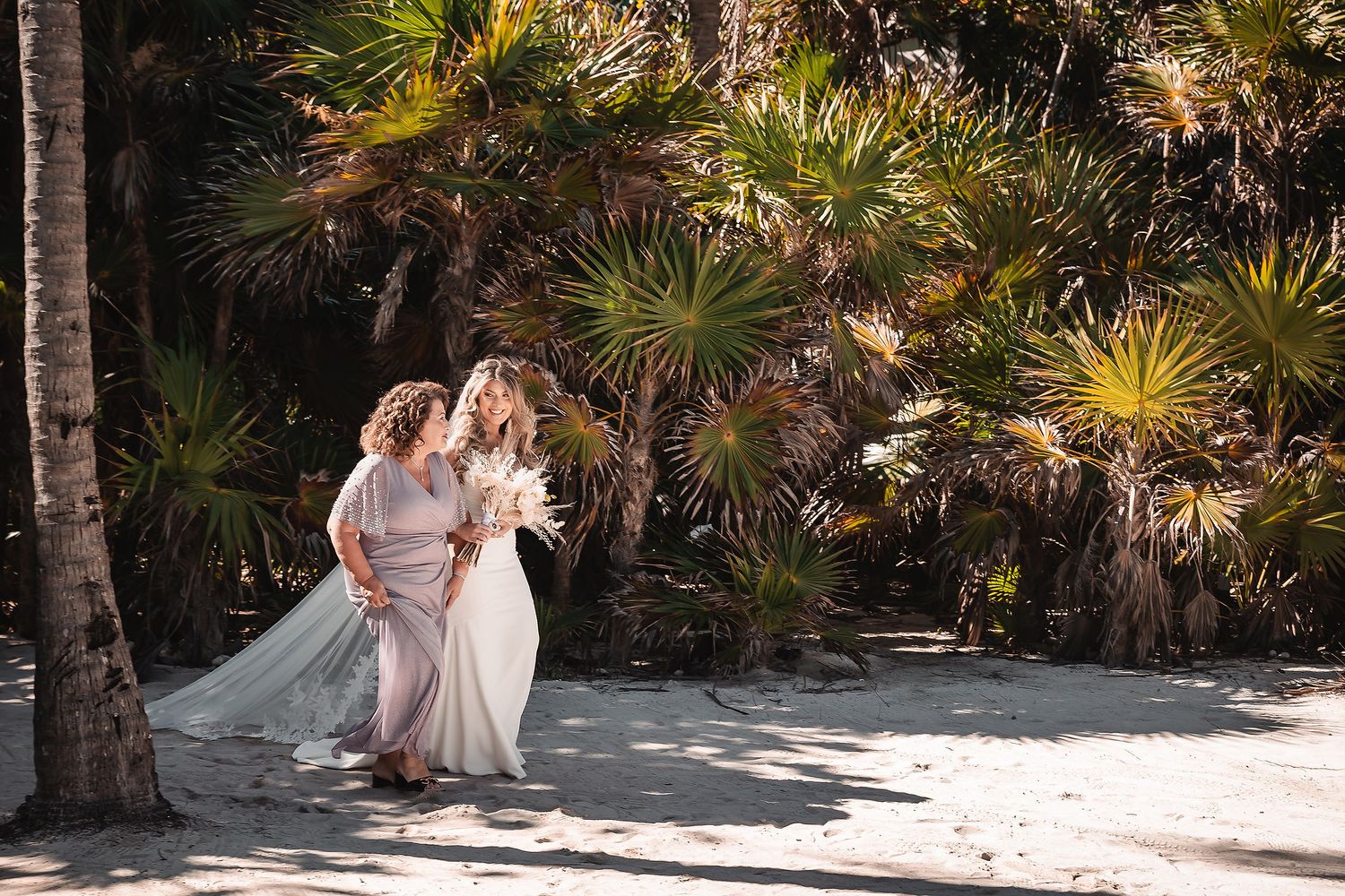 A couple in wedding attire walks hand in hand along a sandy beach path lined with tropical palm trees.