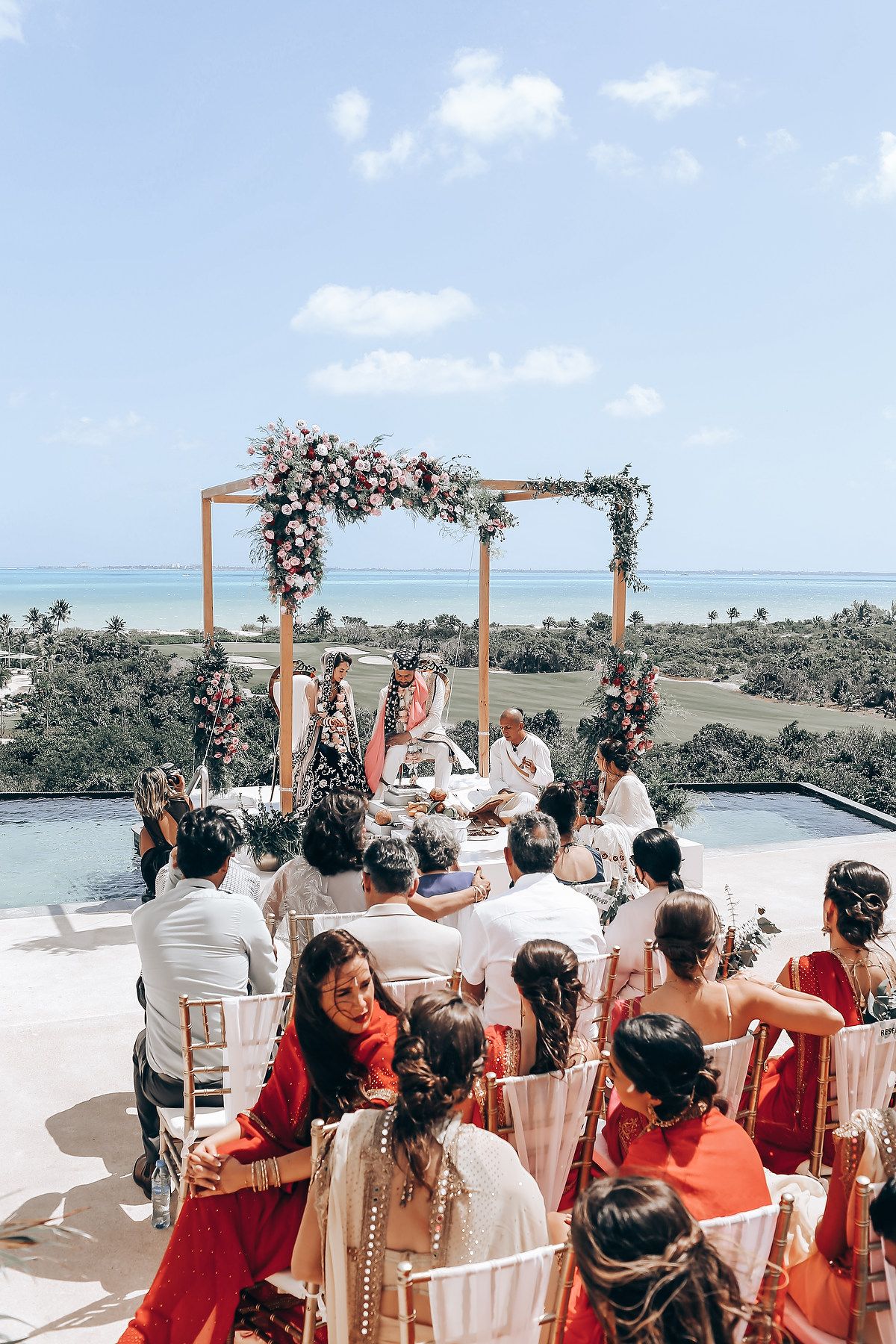 Guests in colorful attire attend an oceanfront wedding ceremony under a floral arch.