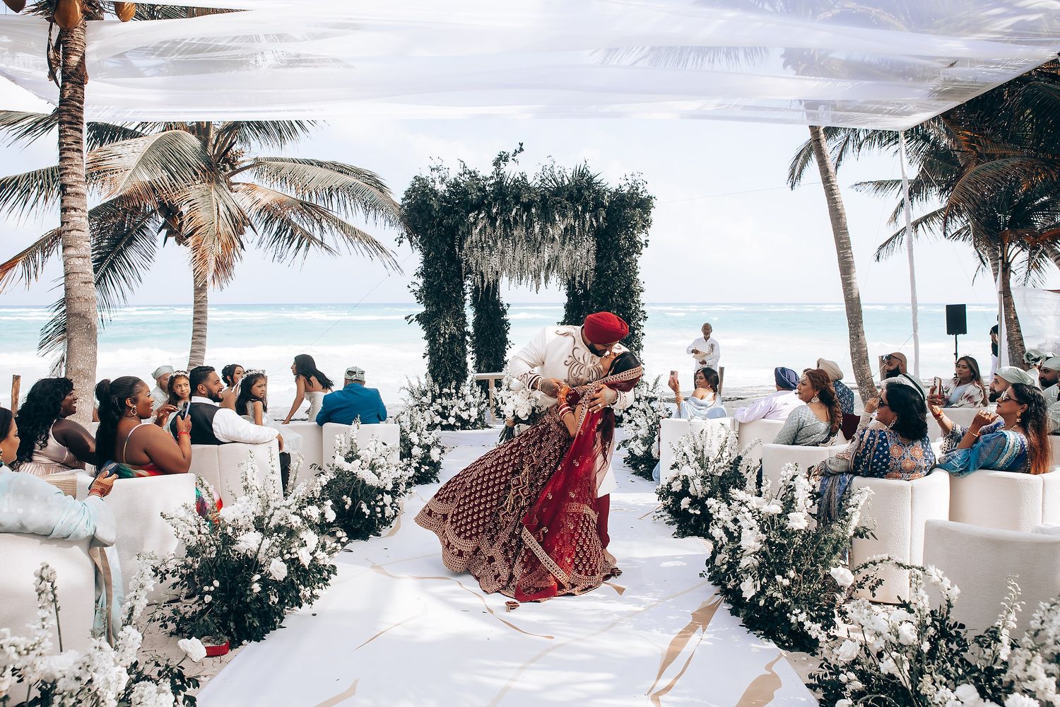 Scenic beach wedding ceremony with guests seated under palm trees.