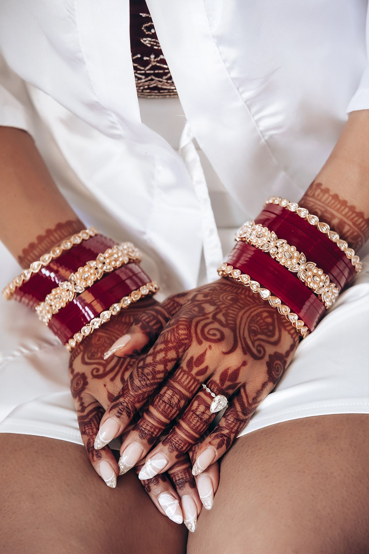 Hands adorned with intricate henna designs and red and gold bangles against a white dress background.