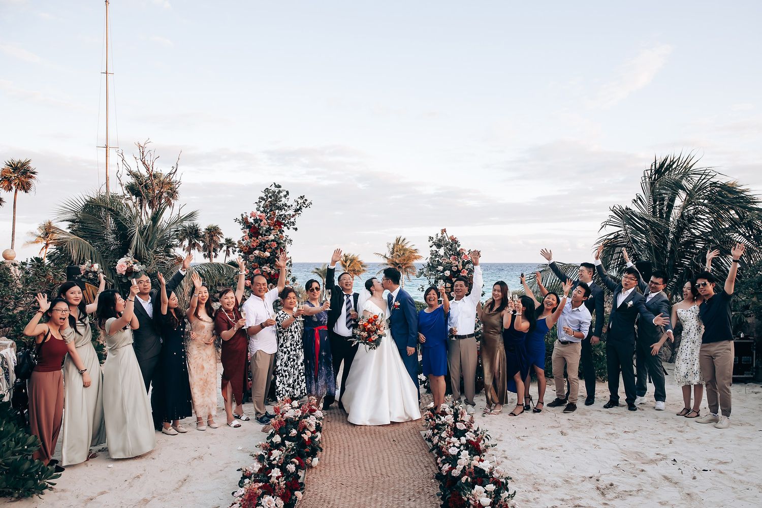 Panoramic view of beach wedding celebration with guests gathered around floral-decorated ceremony space.
