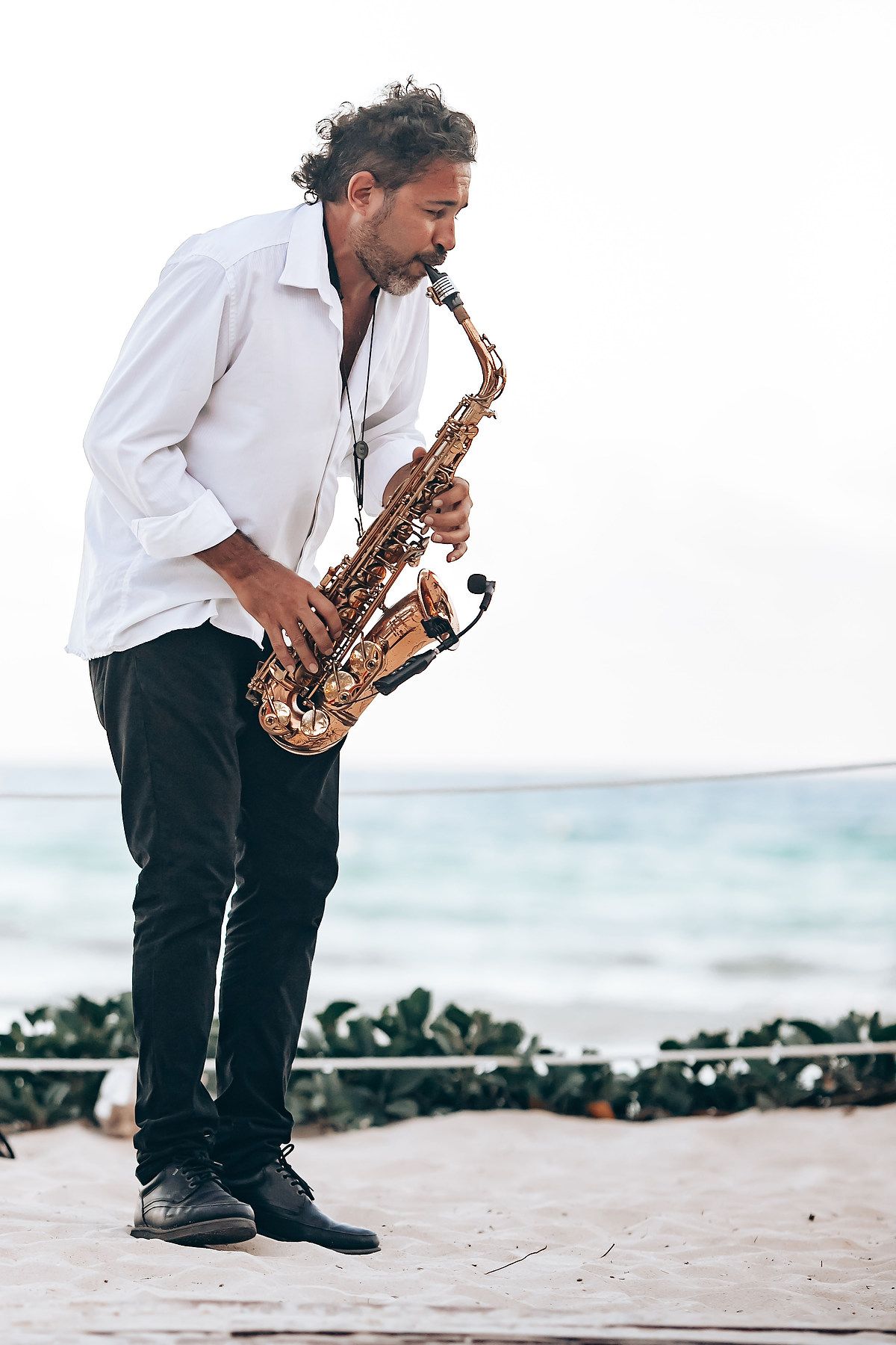 Musician playing saxophone on a beach at sunset wearing formal attire.