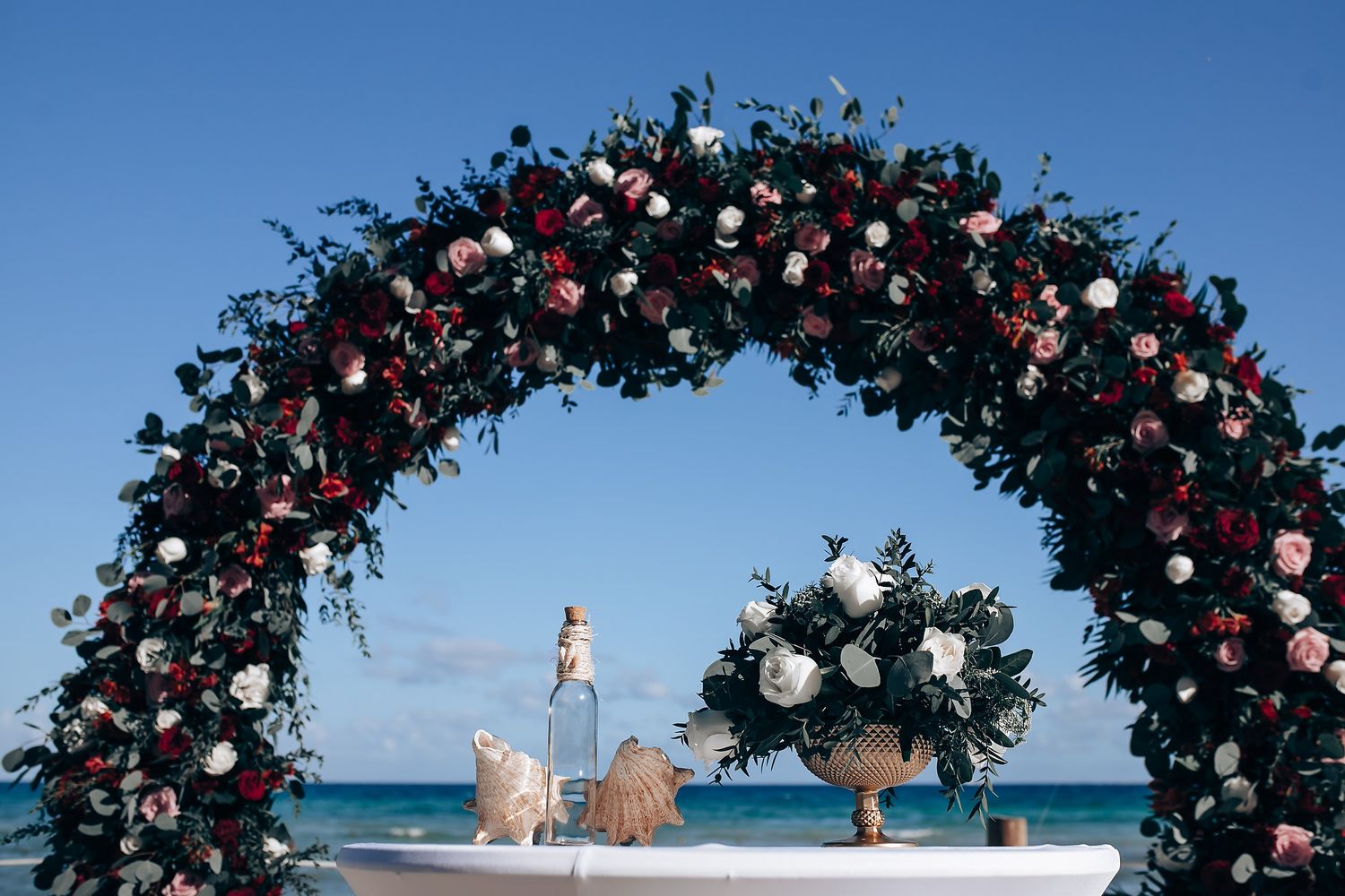 Floral wedding arch with pink and white roses frames a stunning beachfront ceremony setup against blue ocean.