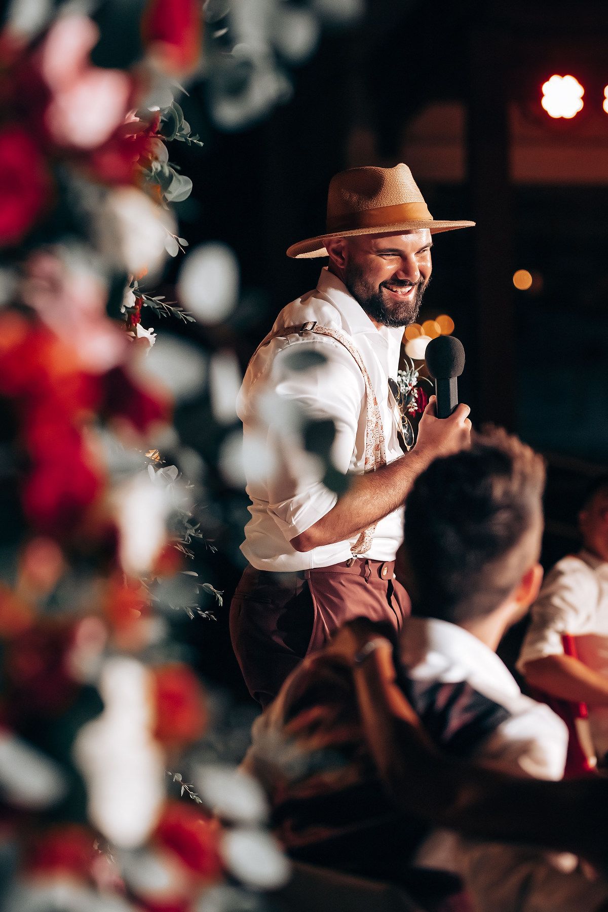 A performer in a brown hat and suspenders sings into a microphone surrounded by floral decorations.