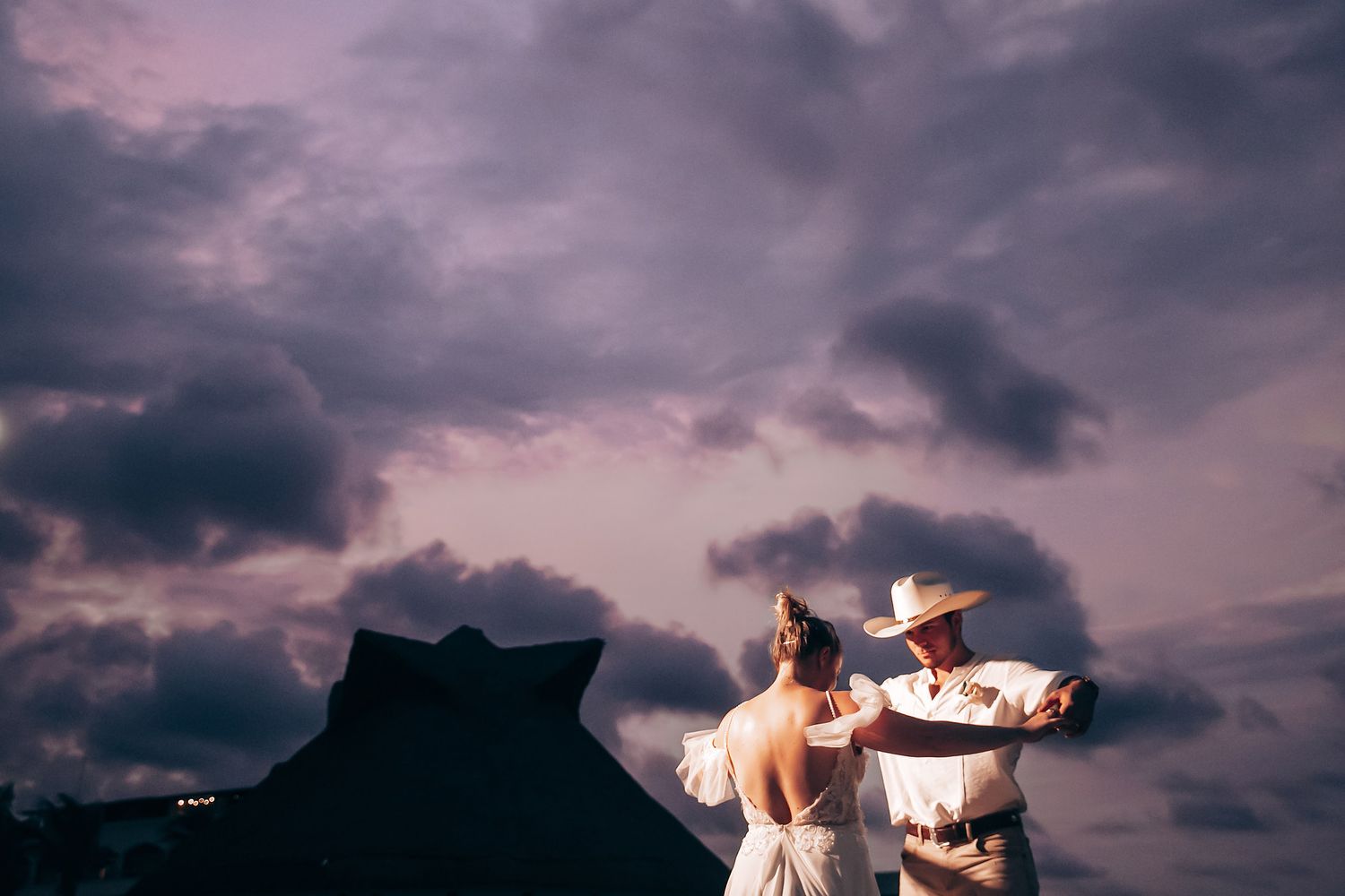 Couple dancing under dramatic purple sunset sky at a beach wedding reception.