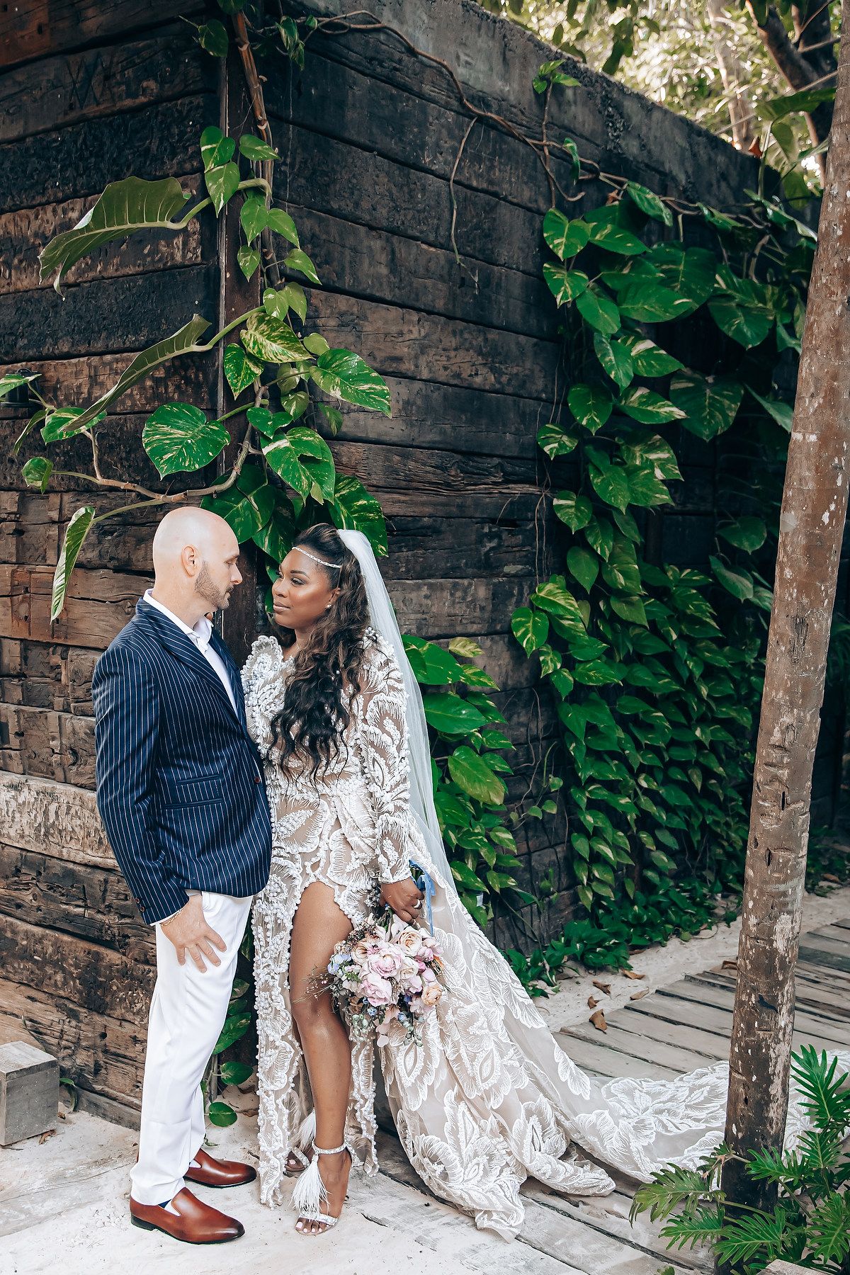 Couple posing against rustic wooden wall with lush green vines for wedding photos.
