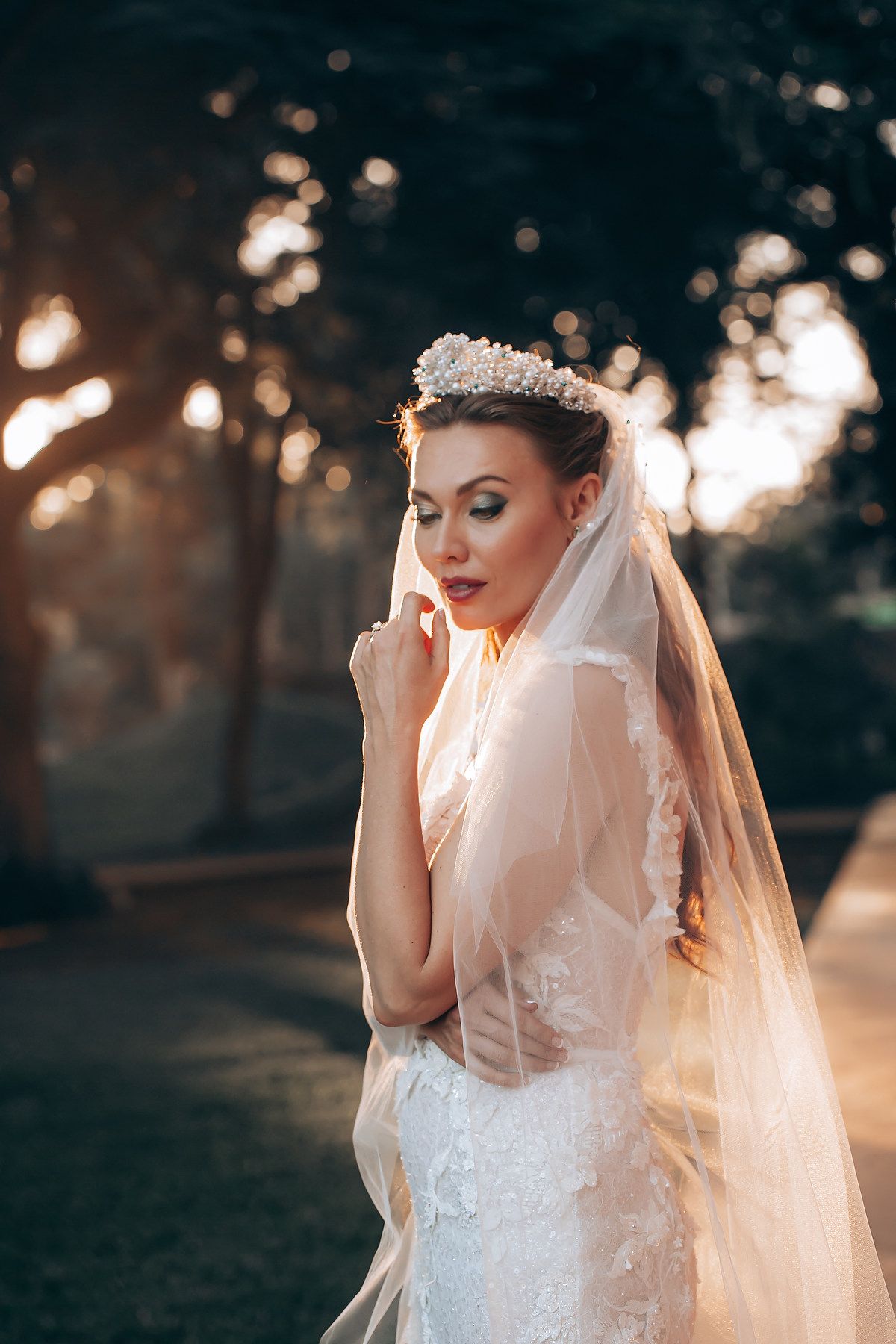 A bride in a white lace gown and flowing veil poses in golden sunset light during an outdoor photo session.