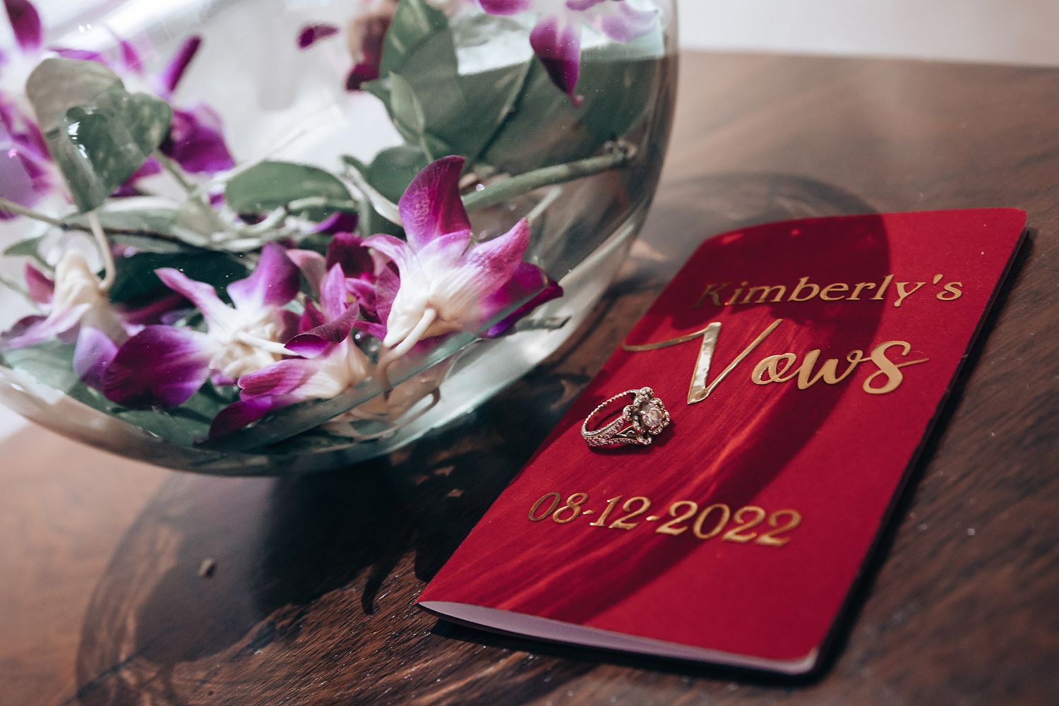 Red wedding vow books decorated with purple orchids and greenery rest on a dark wooden surface.