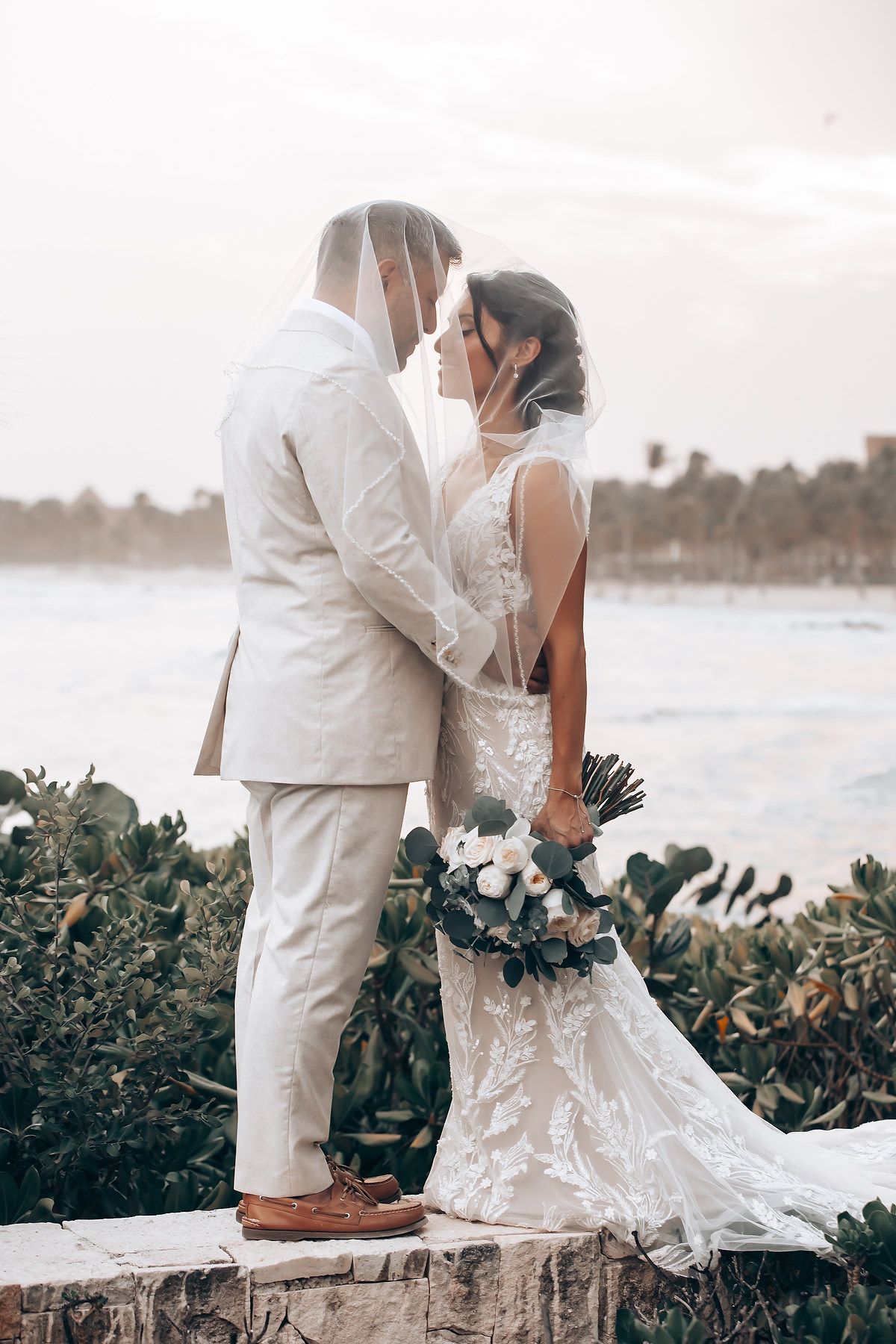 Newlyweds share a romantic moment on a stone wall by the beach at sunset.