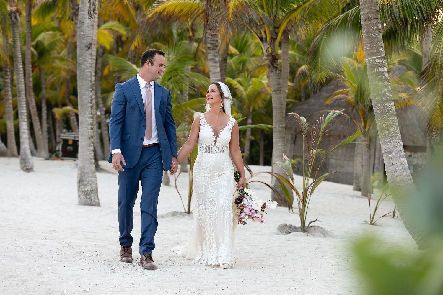 A couple walks hand in hand on a tropical beach with palm trees and white sand in the background.