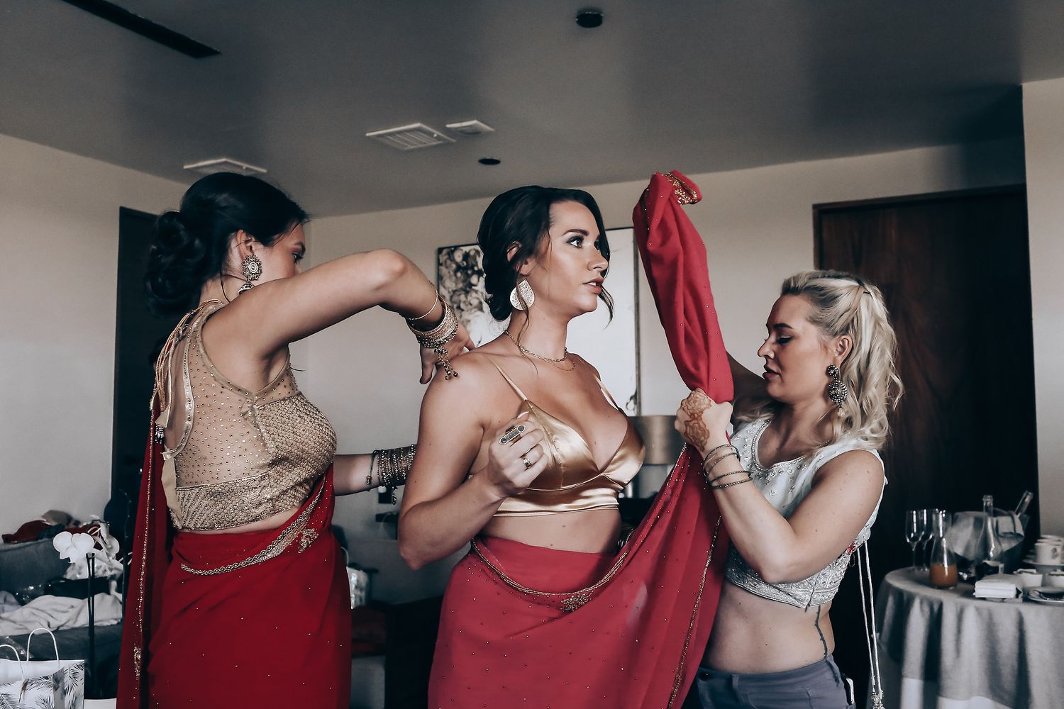 Wedding party members in burgundy dresses help each other get ready in a hotel room.