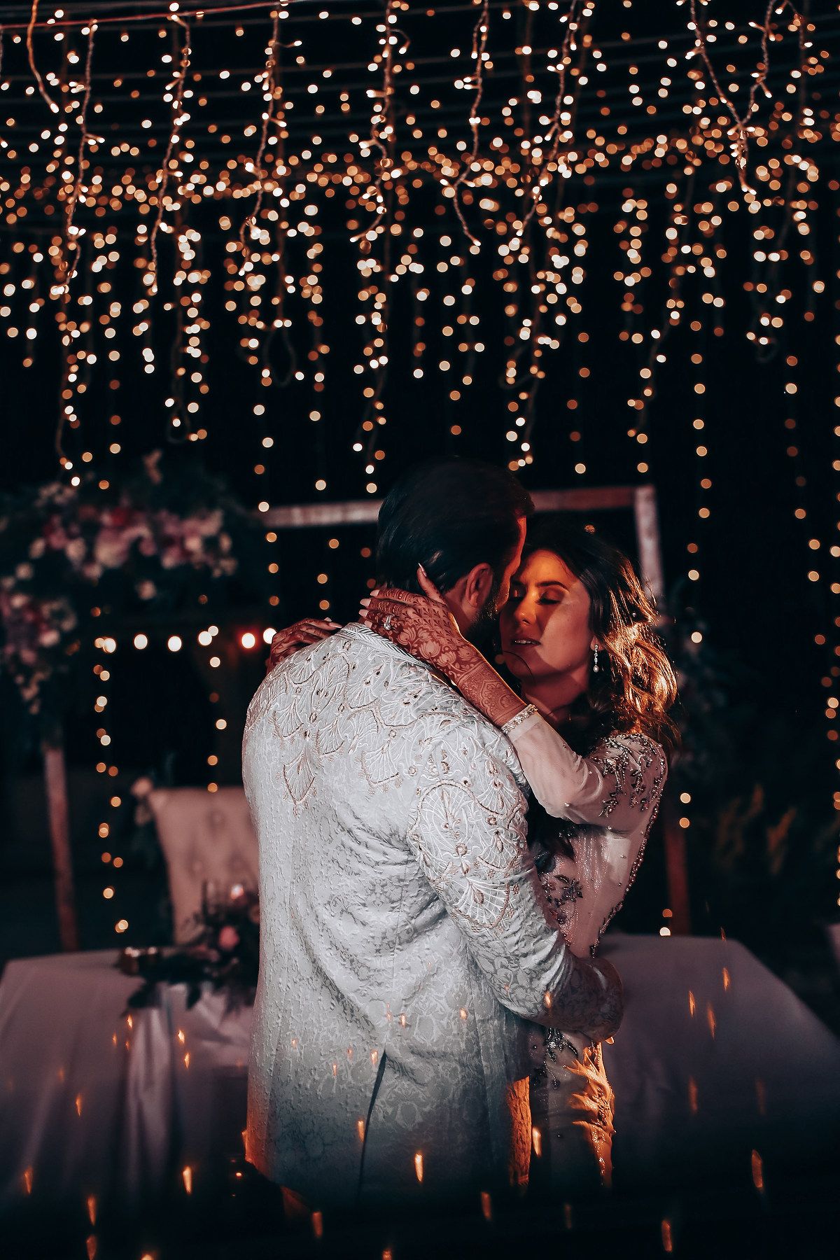 Romantic couple sharing a tender kiss under twinkling fairy lights at an evening wedding reception.