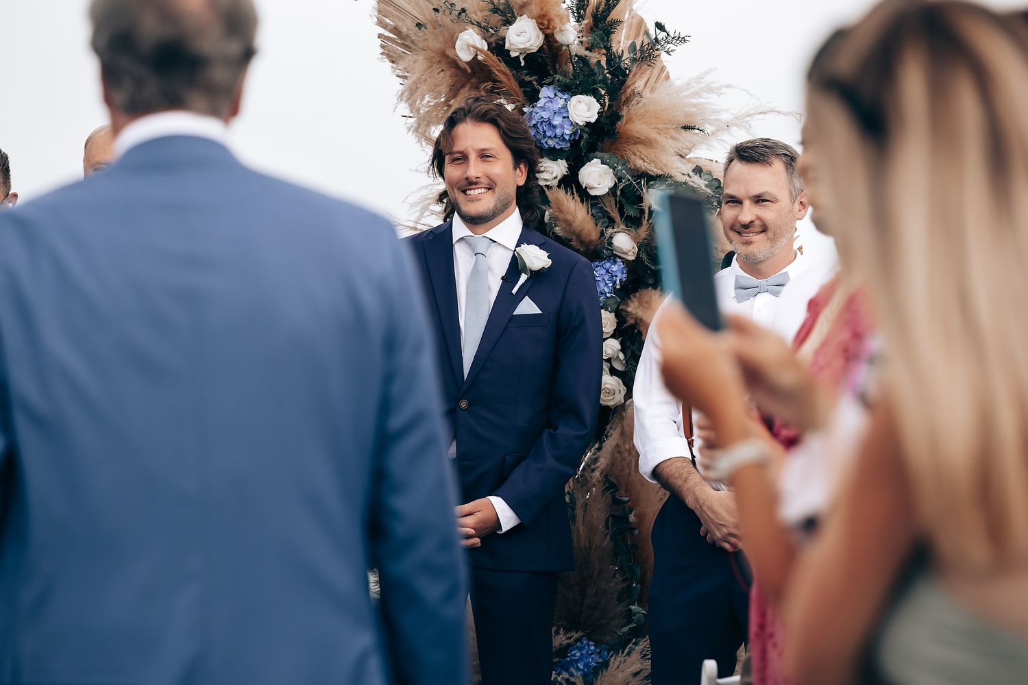 Wedding guests celebrate during an outdoor ceremony with floral arrangements and palm trees.
