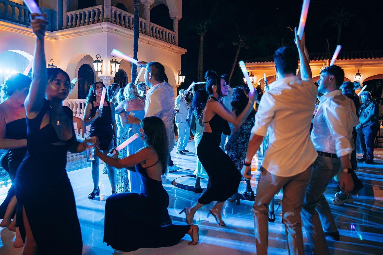 Crowd dancing at a nighttime outdoor party with blue lighting and architectural backdrop.