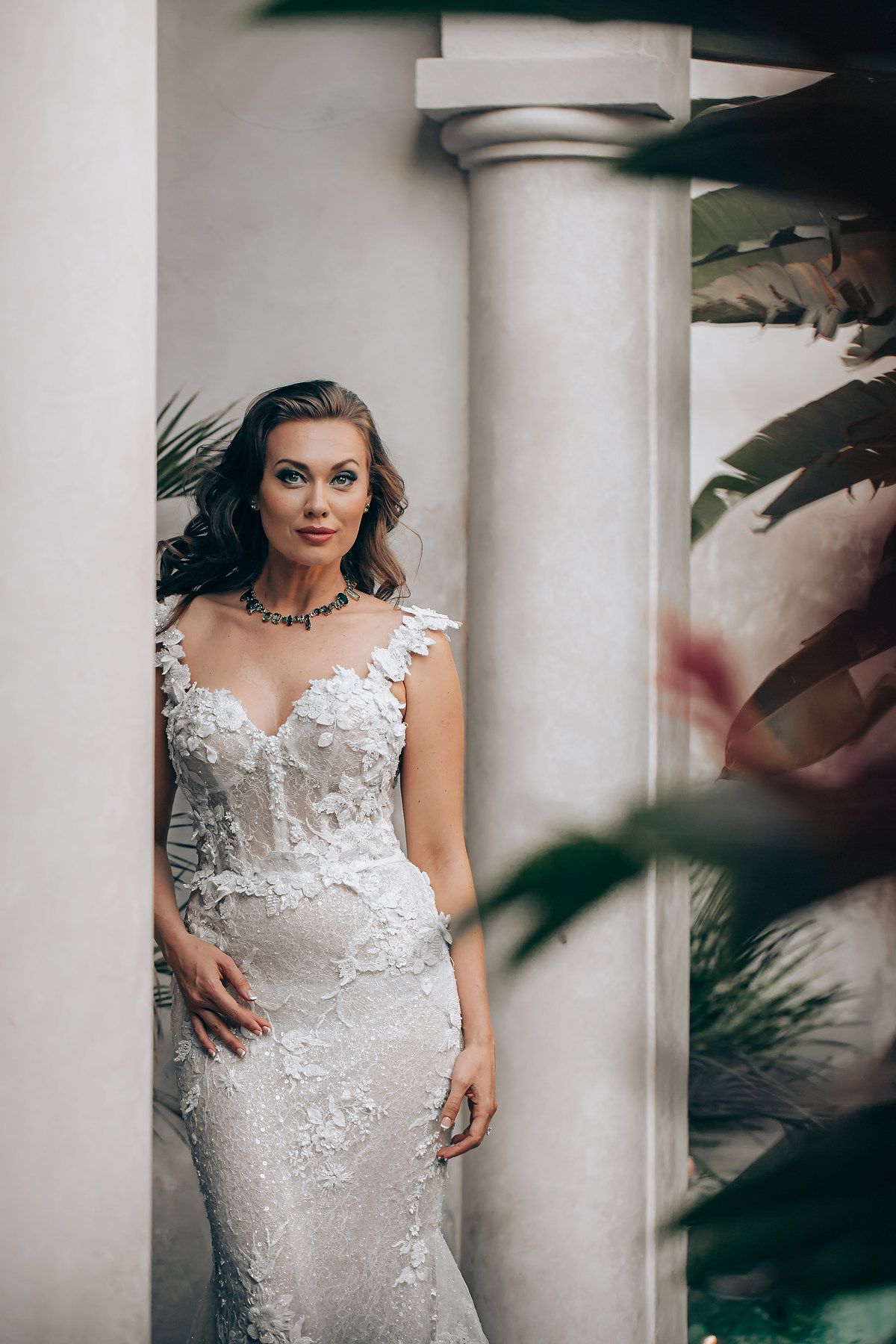 Elegant white lace wedding dress with floral appliques photographed against a classical column and tropical foliage.