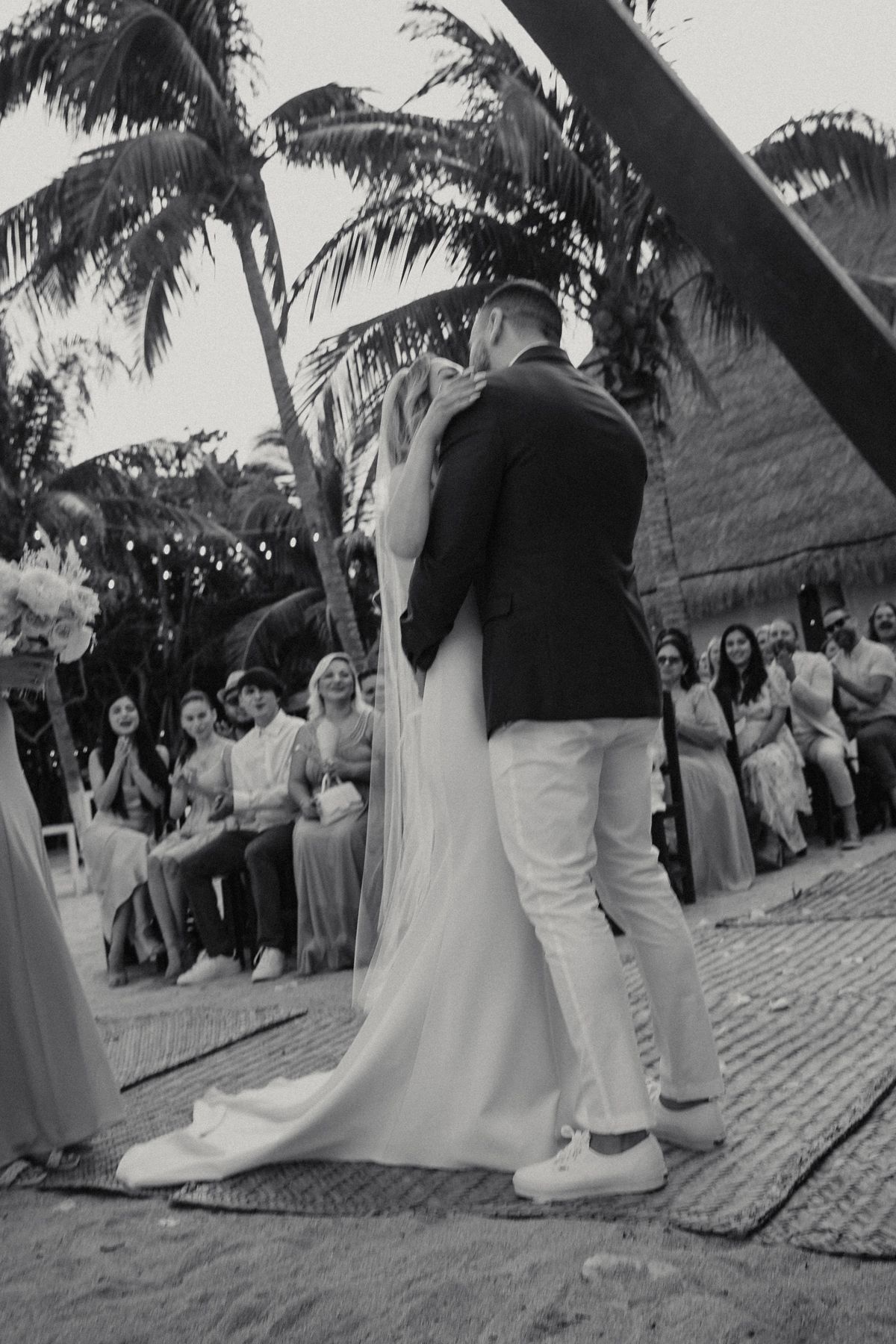 Black and white photo of bride and groom sharing first kiss at tropical beach wedding ceremony.