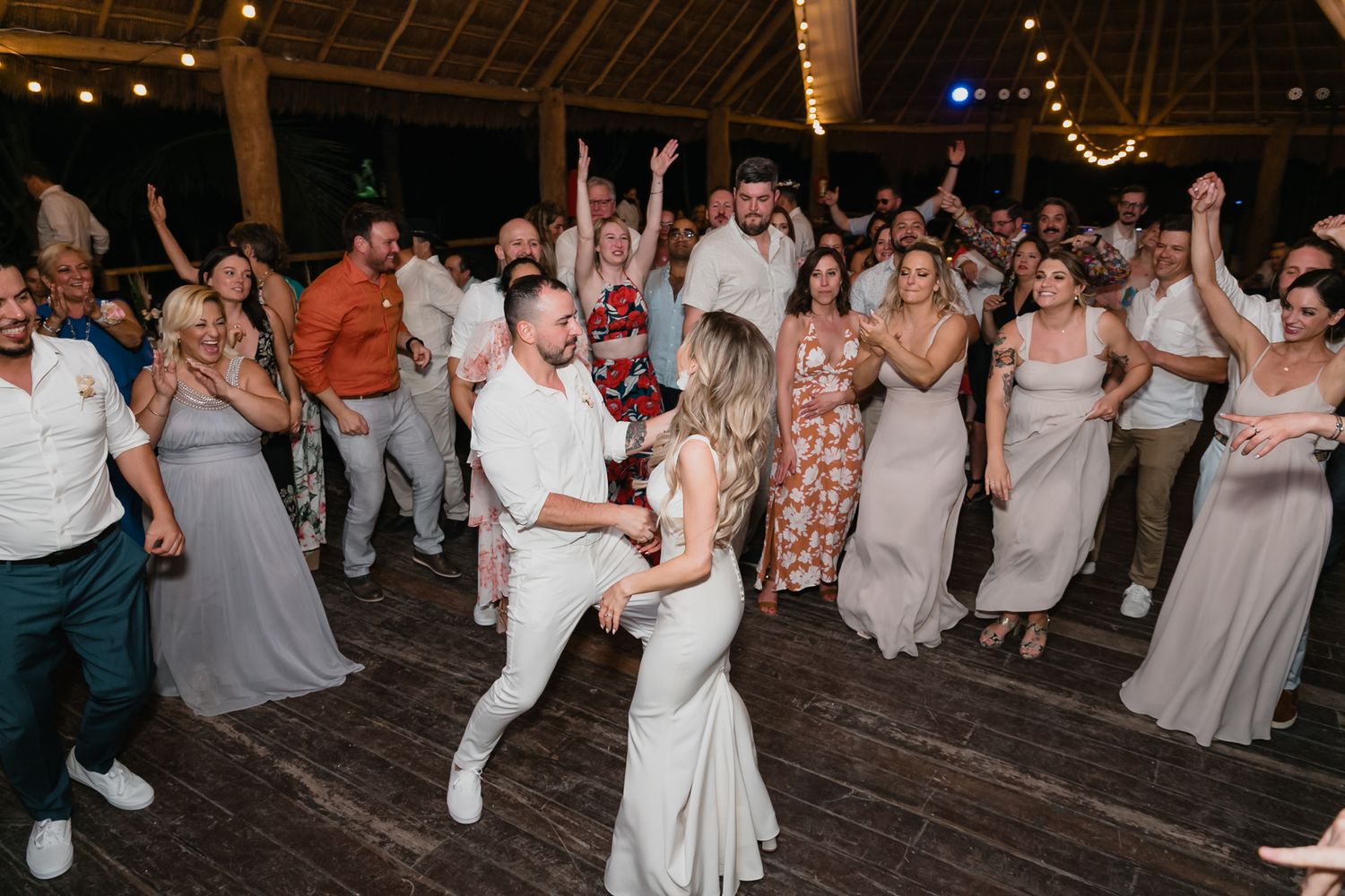 Bride and groom dance joyfully surrounded by celebrating wedding guests on crowded reception floor.
